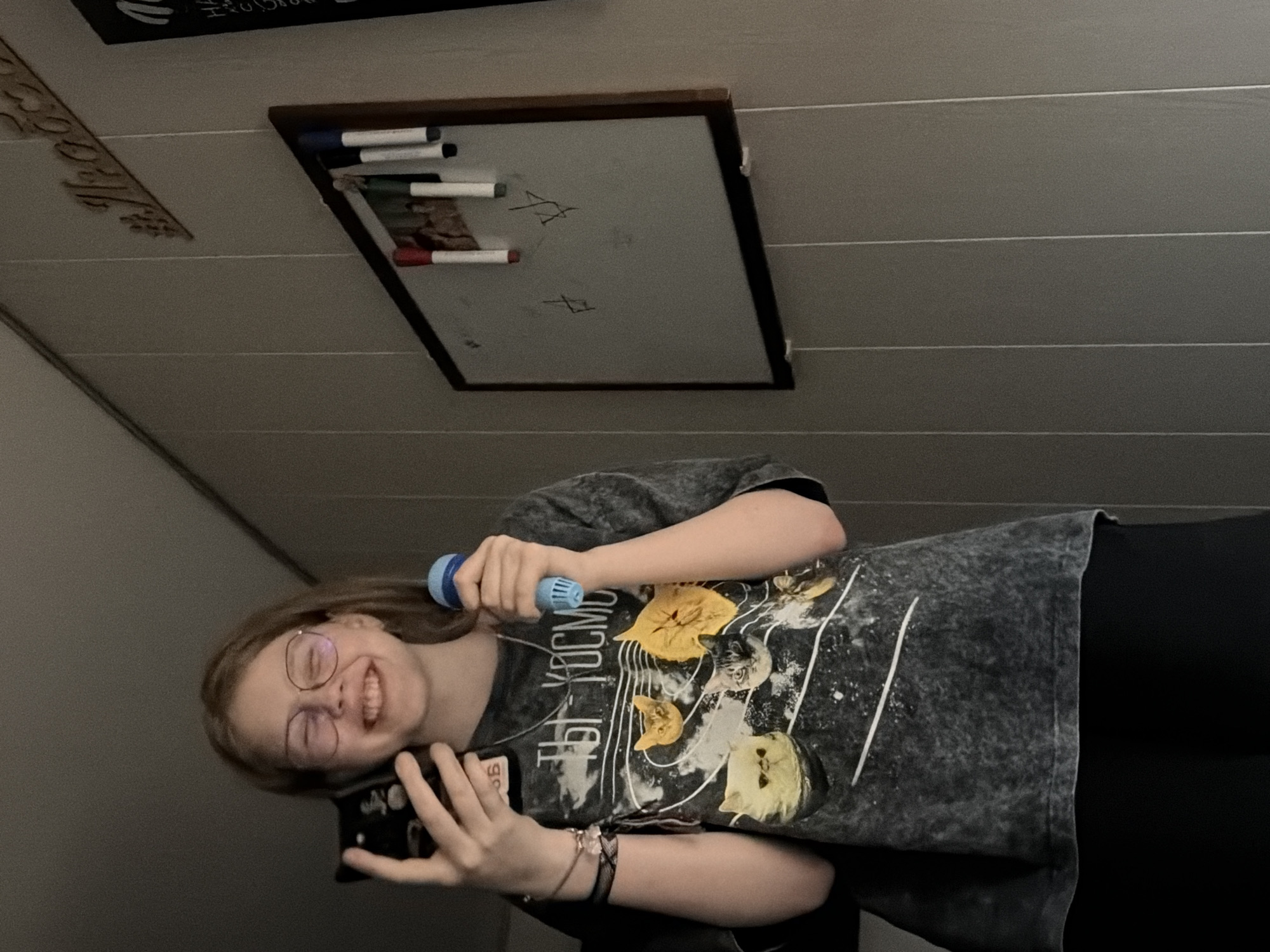 A fair-skinned, smiling person with glasses, holding a black smartphone in their left hand and a blue microphone in their right, is positioned in the foreground of the image. They are wearing a dark-colored t-shirt with a graphic design featuring cats and possibly Cyrillic script. The background consists of a ceiling with horizontal paneling and a framed white board with writing and markers attached. Based on the Cyrillic text potentially present on the shirt, there is a possibility the photo was taken in a location within Russia, such as Chaikovsky.