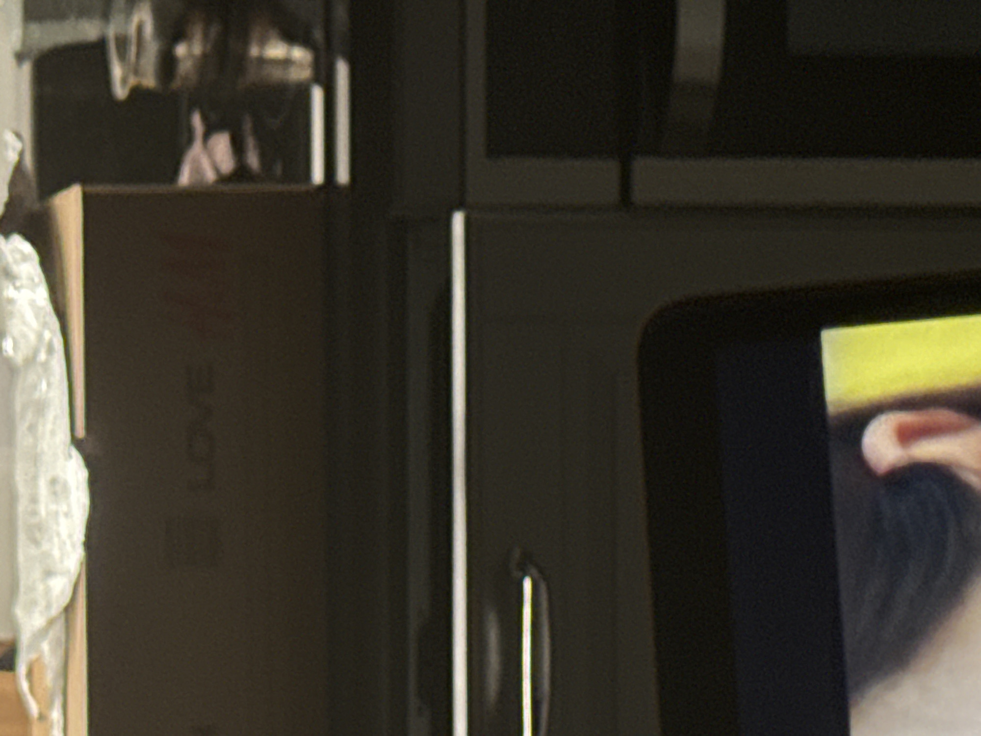 The image shows a close-up view of a dark brown cardboard box with the word "LOVE" printed on it in white. A crumpled piece of silver plastic wrap is draped over the top edge of the box. To the right of the box, a dark, reflective surface is visible, possibly a countertop or shelf, with a blurred object in the background that appears to be a glass jar with liquid inside. Below the box, a dark drawer with a silver handle is partially visible, suggesting this might be furniture. In the lower portion of the image, a person's face is visible, specifically their ear and hair, against a bright yellow background. The scene appears to be indoors, with artificial lighting.