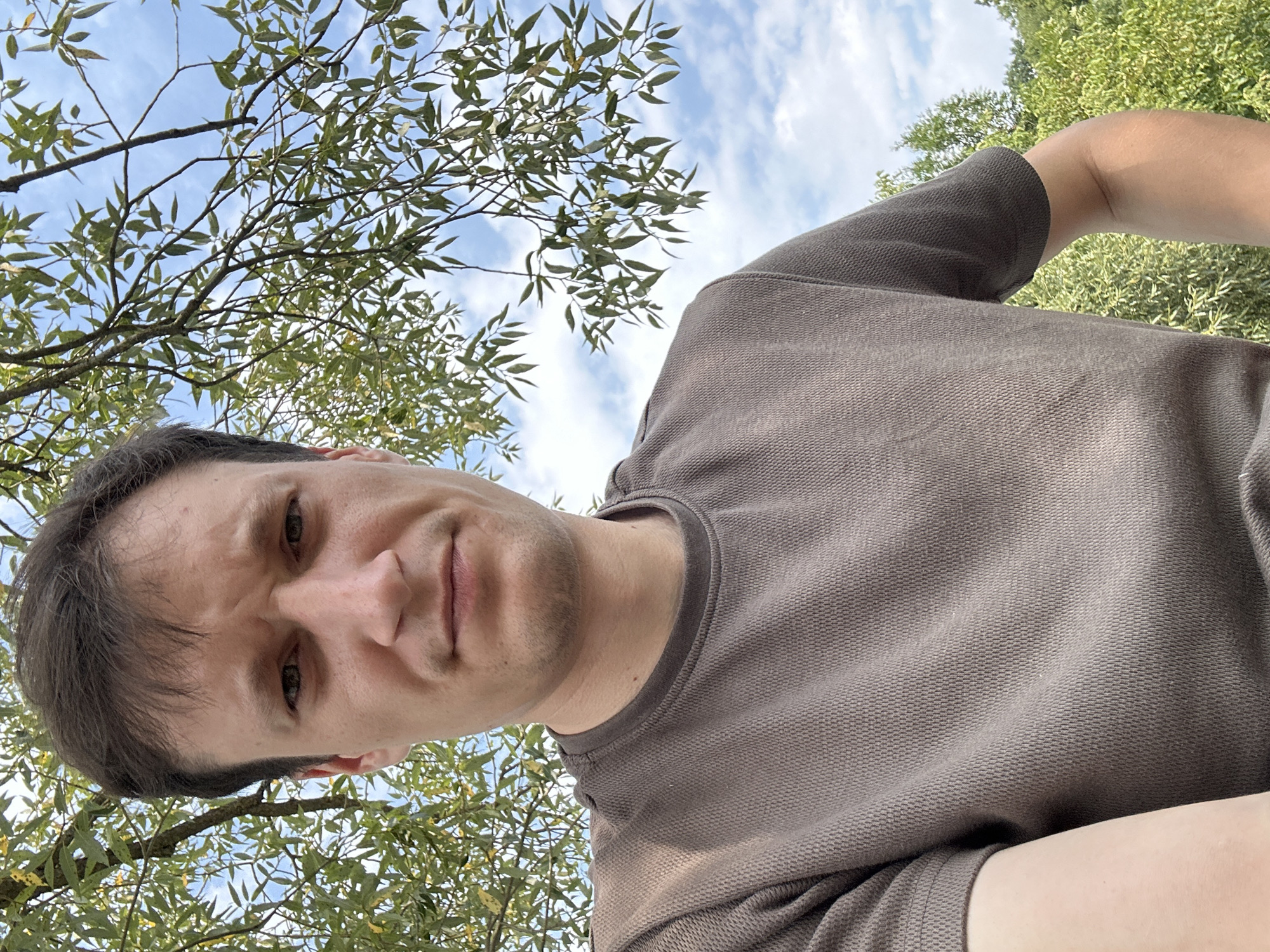 A close-up, low-angle selfie is taken by a man outdoors, with a focus on his face and upper body. He is wearing a brown t-shirt with a textured knit. His hair is dark brown and appears slightly disheveled. His expression is neutral, with his eyebrows slightly furrowed. The background shows a bright blue sky with scattered white clouds, and green foliage from tree branches hanging down. The lighting suggests it is daytime, possibly late morning or early afternoon. The image does not contain any information to determine the specific location within Makiv, Ukraine.