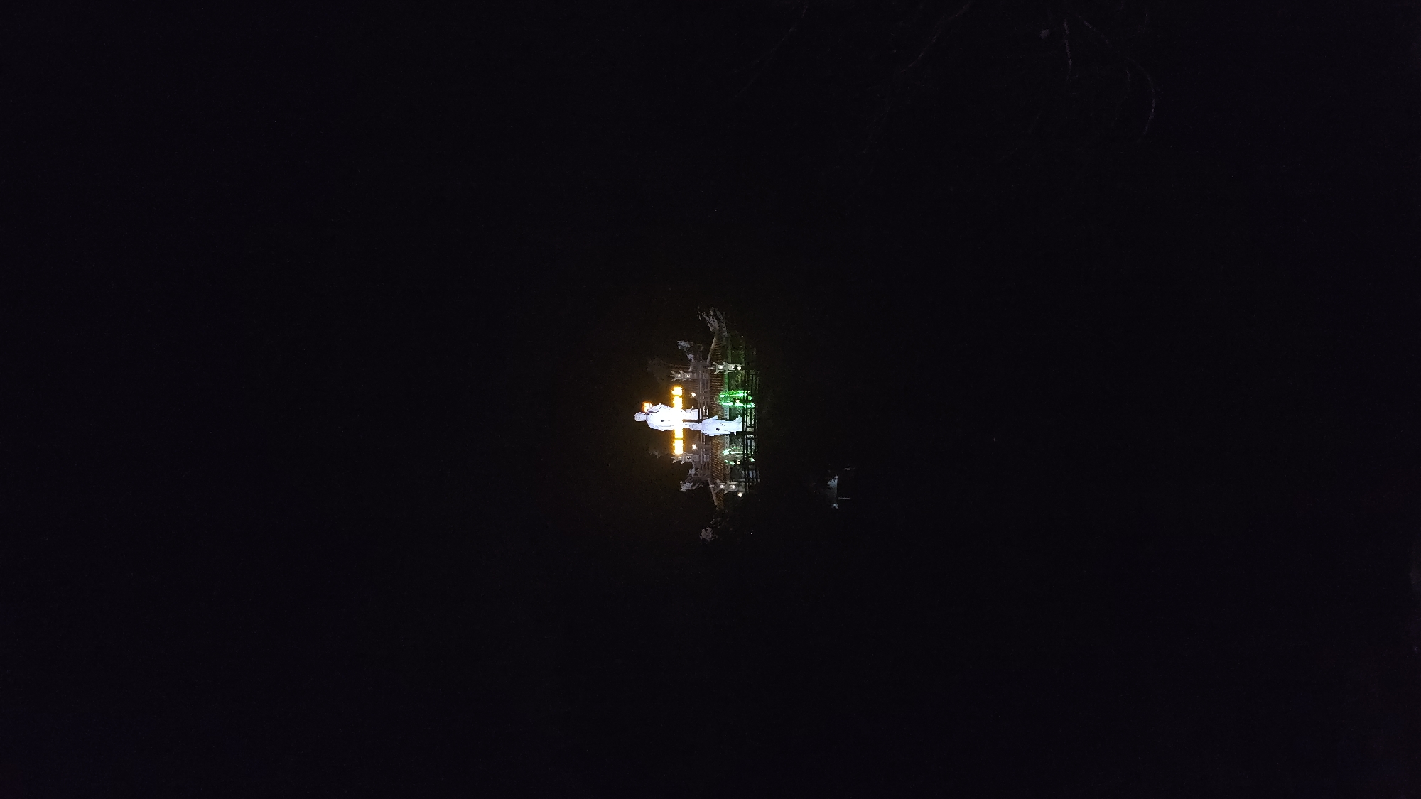 In the center of the image, in Suối Dầu, Vietnam, is an illuminated structure at night. The structure appears to be a monument or statue complex with both white and green lighting. Its reflection is visible in what seems to be water or a dark, reflective surface. The surrounding environment is predominantly dark, suggesting a nighttime setting.