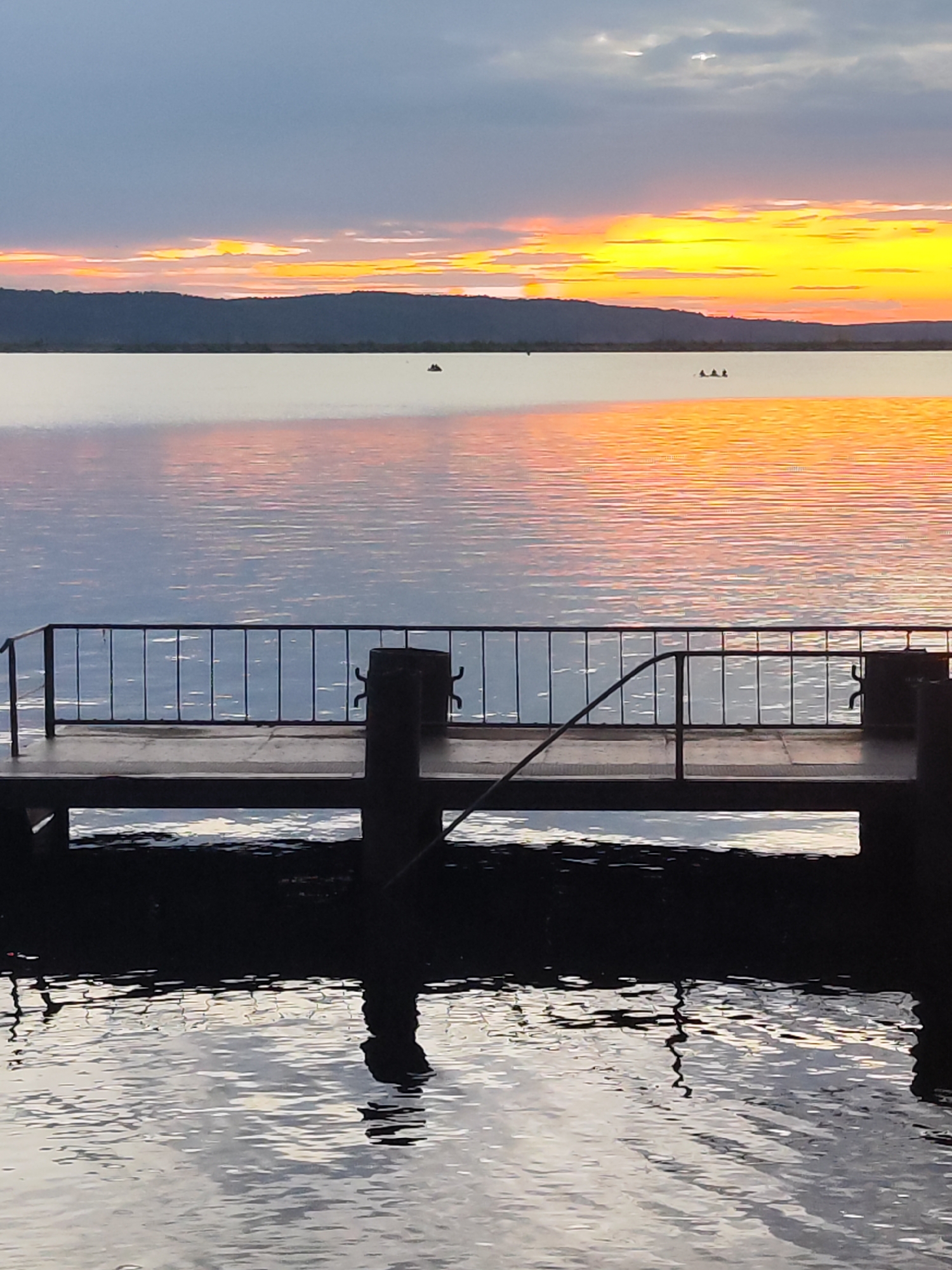 The image depicts a sunset over a body of water, likely a river or lake, with a dock or pier in the foreground. The sky is filled with vibrant colors of orange, yellow, and pink, reflecting on the calm water. A dark silhouette of a treeline or hills is visible in the distance, separating the water from the sky. On the pier, there are metal railings and posts. In the water, a few small objects, possibly boats or buoys, are visible. The overall scene is serene and picturesque. The location is the Kama River in Chaikovsky, Russia, known for its scenic beauty, especially during sunset.