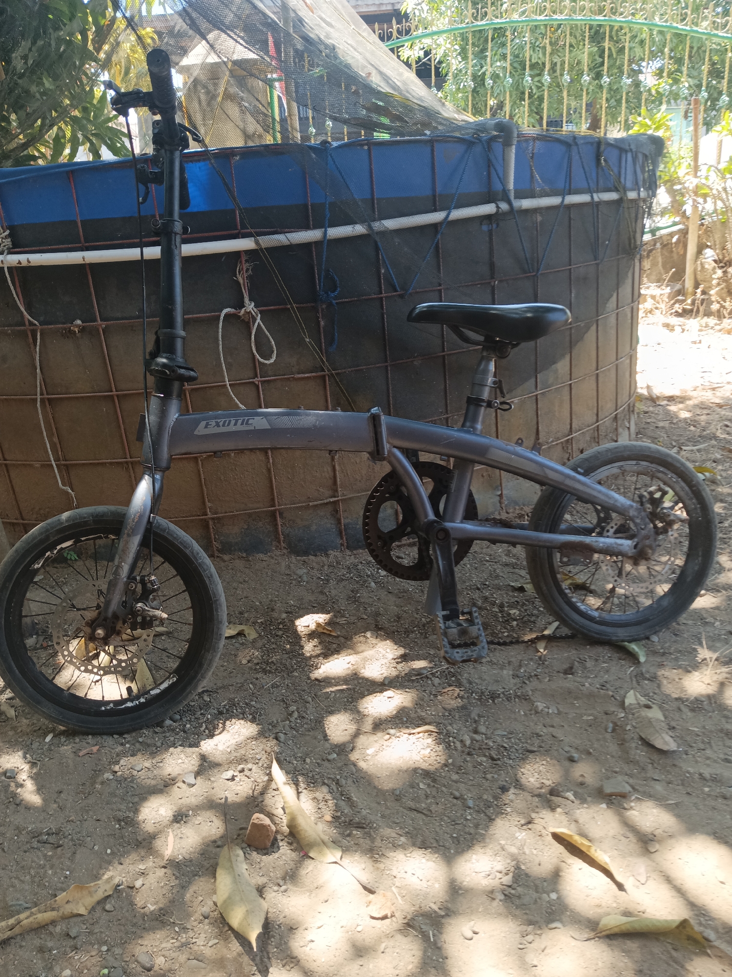 A dark gray folding bicycle with "EXOTIC" branding on the frame is parked on a dirt surface. The bicycle has small wheels with disc brakes, a black seat, and black pedals. It is positioned in front of a large, cylindrical structure made of wire mesh and covered with blue fabric and a black net. The background shows greenery and a light-colored fence. The scene appears to be outdoors, with shadows indicating it is daytime. The overall environment suggests a somewhat rural or undeveloped area, possibly a yard or open space in a village like Pitue, Indonesia. There are no people visible in the image.