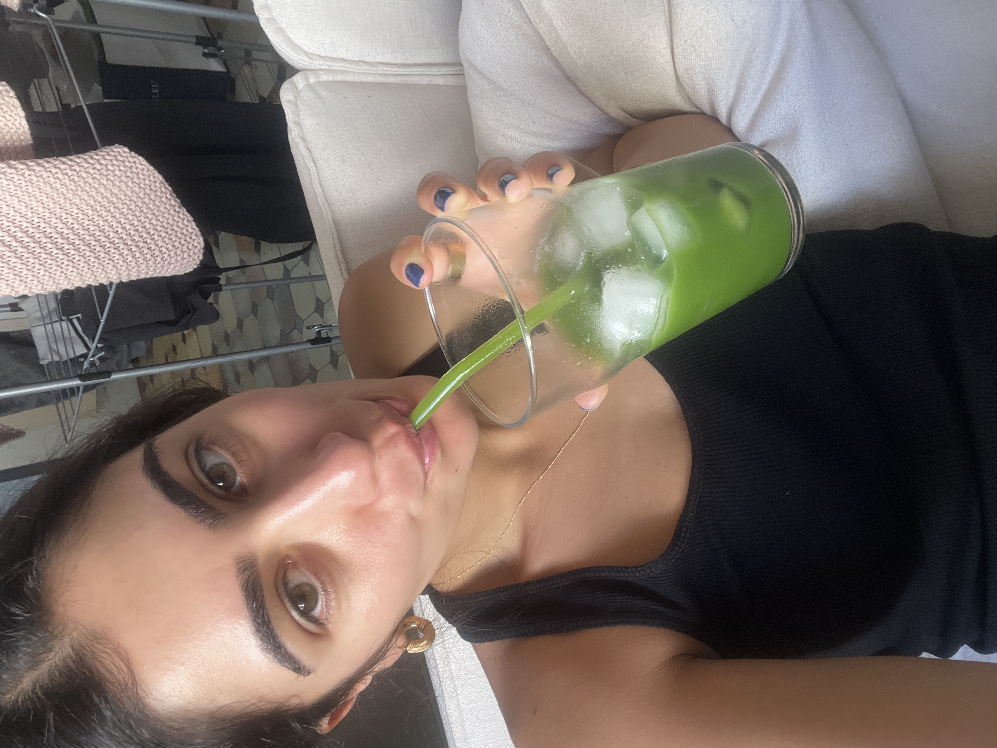 A woman with dark hair and light brown eyes is drinking a green beverage with ice from a clear glass with a green straw. She is wearing a black tank top and a gold hoop earring. The background shows a clothes rack with clothing, a pink knitted item, and white furniture. This appears to be a residential setting, possibly an apartment. The image is a selfie taken from the woman's perspective.