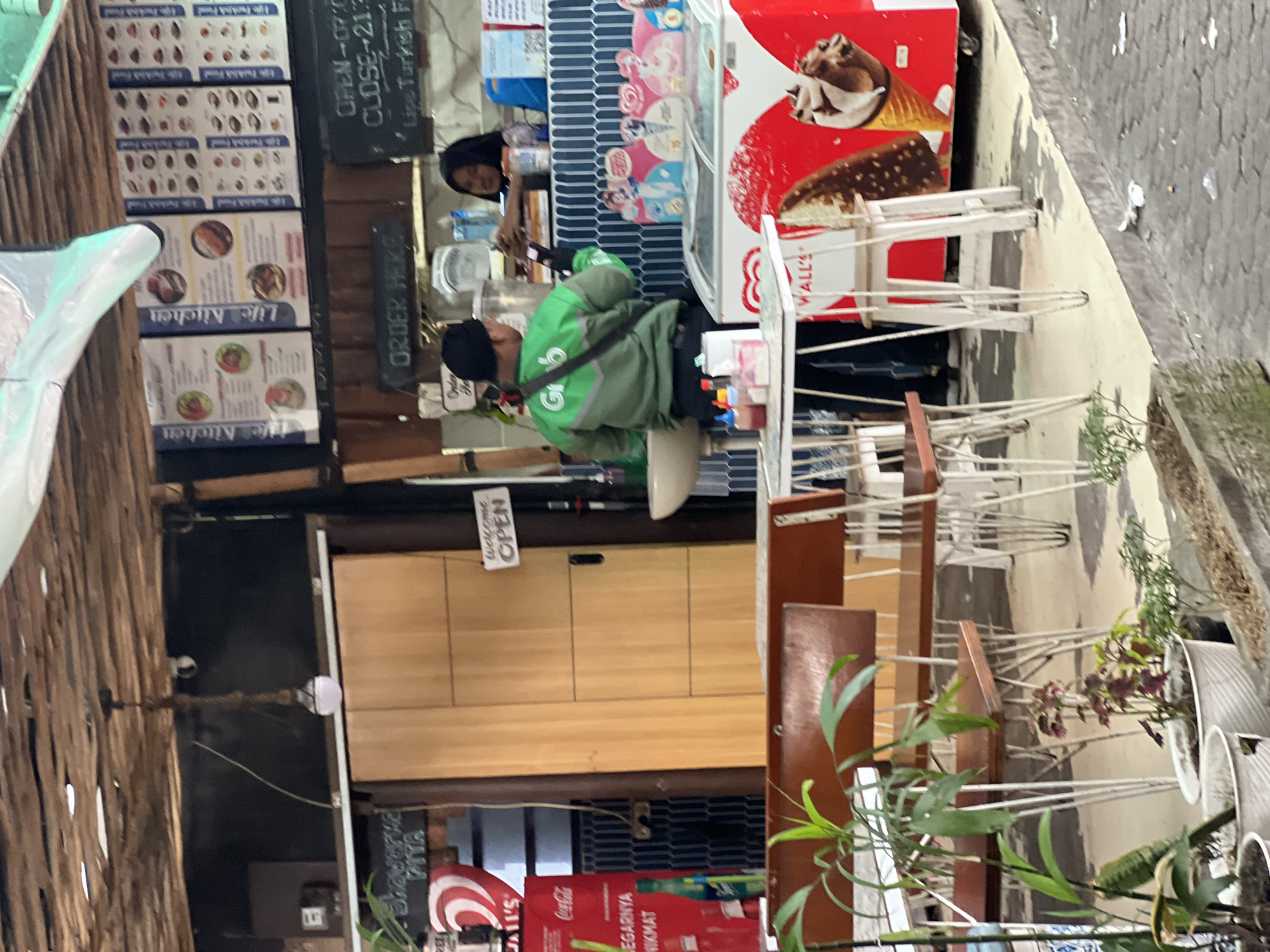 The image shows an outdoor seating area and food stall in Kuta, Indonesia. A delivery driver in a green Grab uniform is handing something to a woman behind the counter, who is wearing a patterned top. To the left, a red sign for Coca-Cola is visible, along with a menu board for "Life Kitchen." An ice cream freezer is prominently displayed with images of ice cream cones. Tables and chairs are set up for customers, and potted plants add greenery to the scene. The overall environment suggests a casual dining establishment.