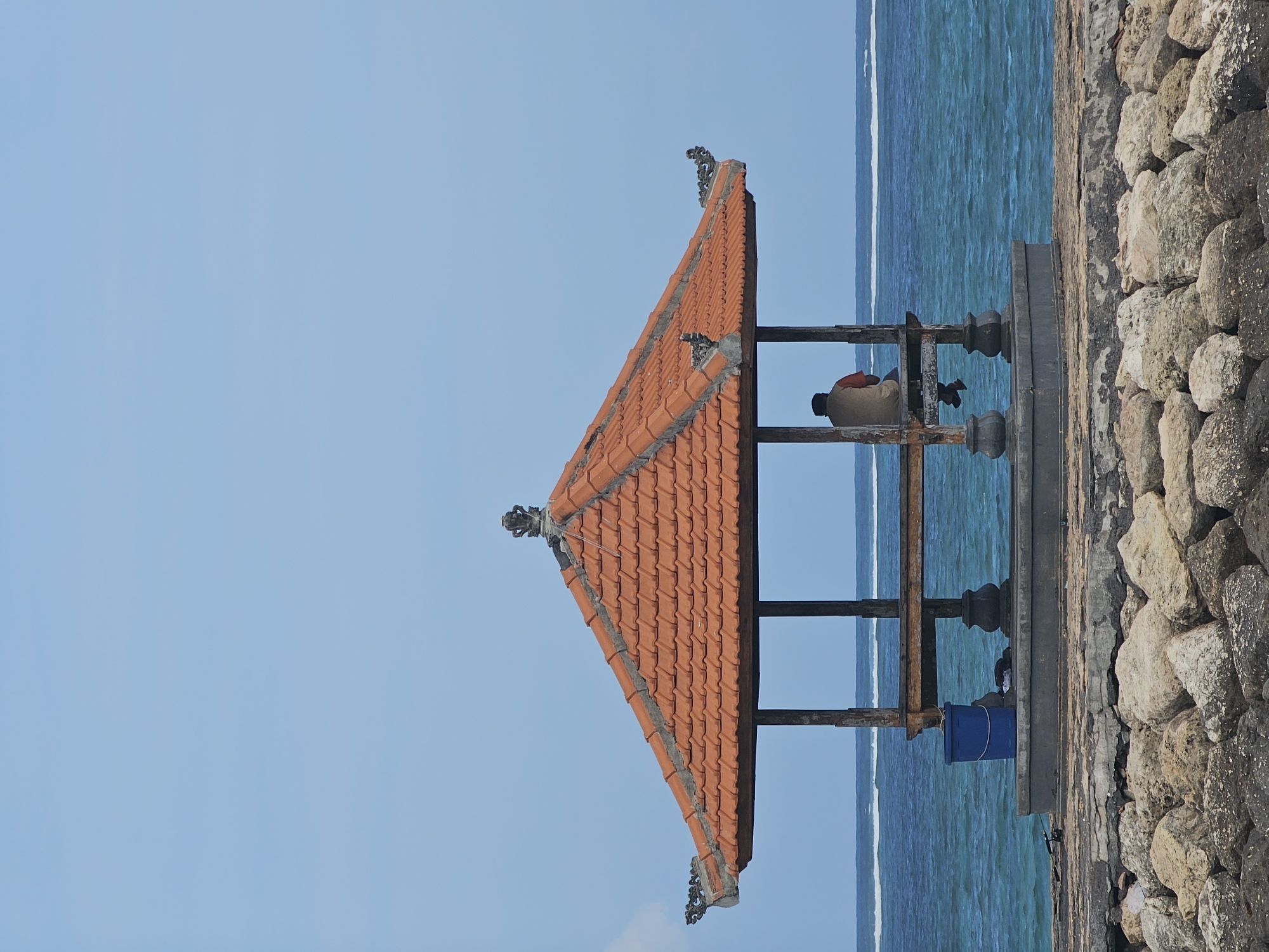 A solitary individual sits within a traditional Balinese-style pavilion with an orange tile roof, overlooking the ocean. The pavilion is supported by dark pillars resting on a concrete platform. In front of the pavilion, a blue bucket is visible. The background features the turquoise ocean with white waves and a clear blue sky. The foreground is a rocky embankment. This scene likely depicts a tranquil moment at a beach or coastal area in Denpasar, Indonesia, a city known for its Hindu temples and beaches.