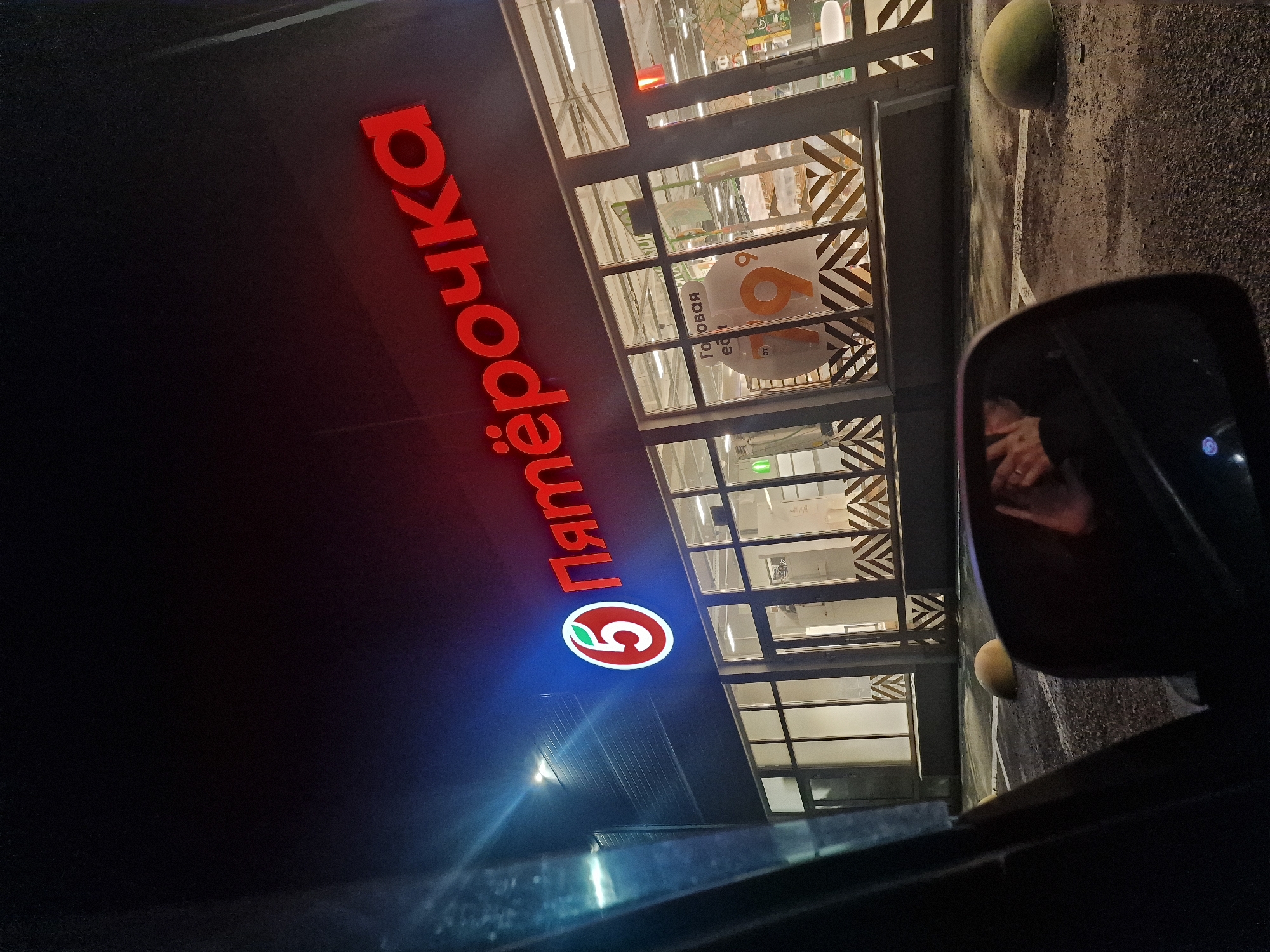 The image shows a nighttime view of the exterior of a "Pyaterochka" (Пятёрочка) grocery store in Kursavka, Russia. The store's prominent red neon sign, reading "Пятёрочка" and featuring the brand's logo of a red circle with a white "5" and a green leaf, is illuminated. The building has large glass windows, some of which are decorated with a geometric chevron pattern. Inside the store, shelves and lighting are visible. The foreground shows a dark car interior, with the reflection of a person's hands in the side mirror. The exterior of the store is paved, with yellow-green concrete bollards visible in the parking area. The environment is dark, indicating it is nighttime, and the lighting from the store is the primary illumination source.