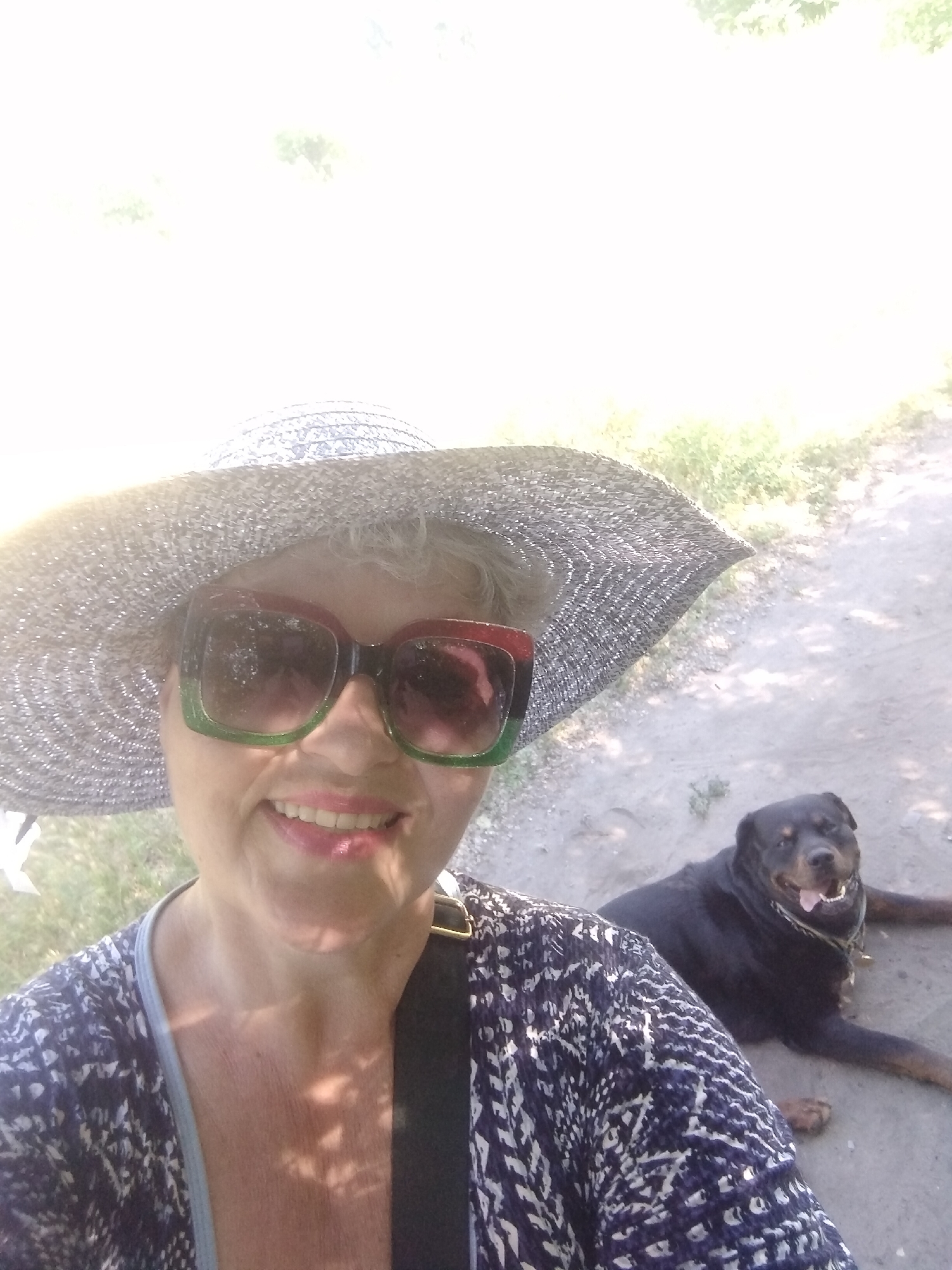 A selfie-style image depicts a woman wearing a wide-brimmed straw hat and large sunglasses, with her dog, a black Rottweiler, lying on the ground behind her. The woman is smiling and appears to be outdoors, with a dirt path and some vegetation in the background. The lighting suggests it is a sunny day. The dog is looking towards the camera with its tongue out.