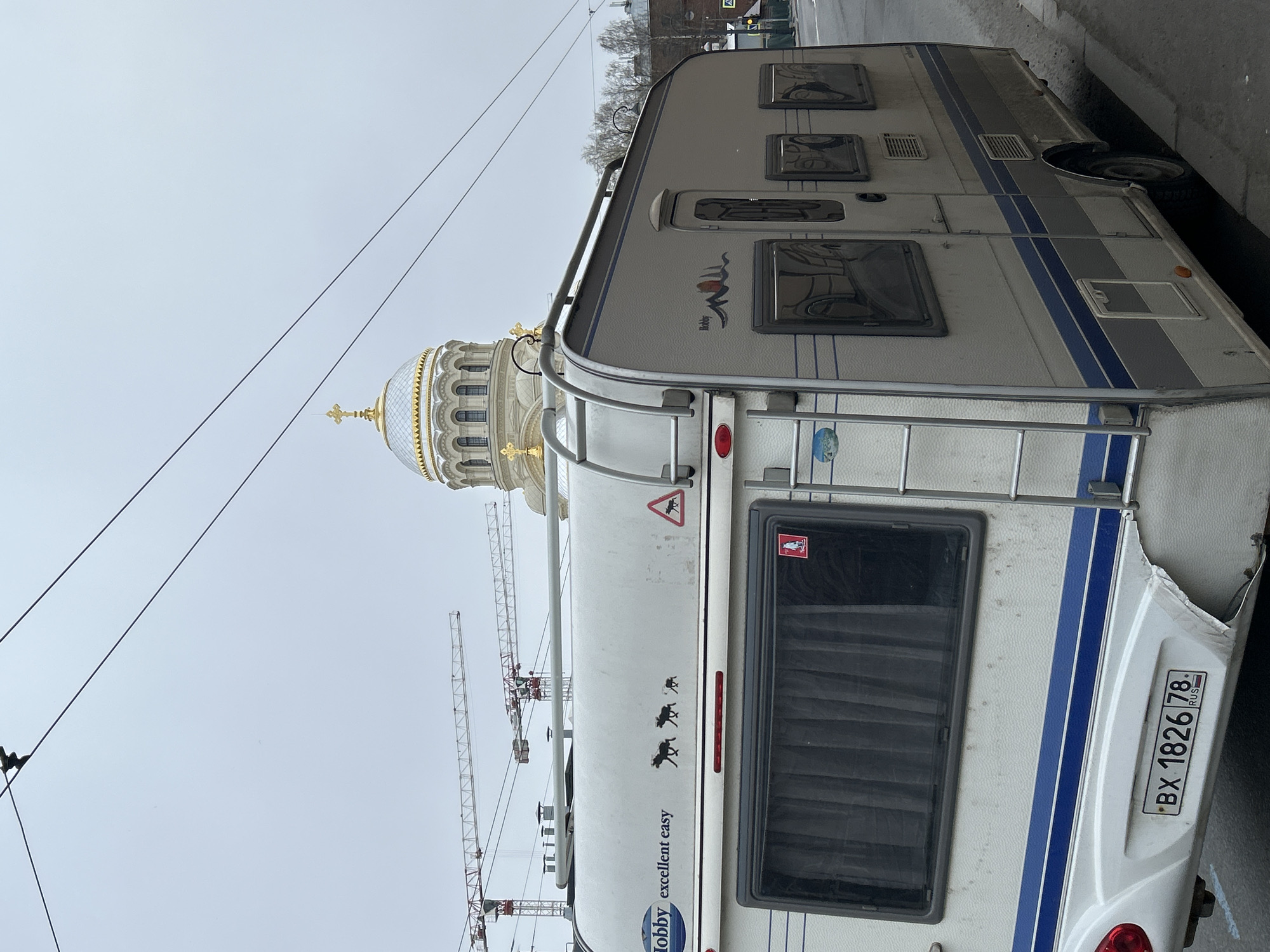 An image captures a white and grey caravan parked on a city street under an overcast sky in Saint Petersburg, Russia. The caravan, seen from its side and rear, dominates the foreground. It features distinct blue stripes along its body, several windows with dark blinds, and roof rails. On its side, the brand name "Hobby" is visible with a logo, and the text "excellent easy" printed below. Several silhouetted decals of moose or deer are affixed to the rear panel, along with a red triangular warning sign depicting an animal, and another red square sticker with a white animal. The rear right corner of the caravan shows some damage. A Russian license plate, "BX 1826 78 RUS", is clearly visible on the back.
In the mid-ground and background, a majestic golden dome of a large, ornate building, likely a cathedral or church, rises prominently, topped with a golden cross. Its classical architecture with rows of arched windows is discernible. Behind the caravan and to the left of the dome, several red and white tower cranes are visible, suggesting ongoing construction in the area. Overhead, multiple power lines crisscross the frame against the pale, overcast sky. The scene depicts an urban setting combining everyday life with historical architecture and modern development.