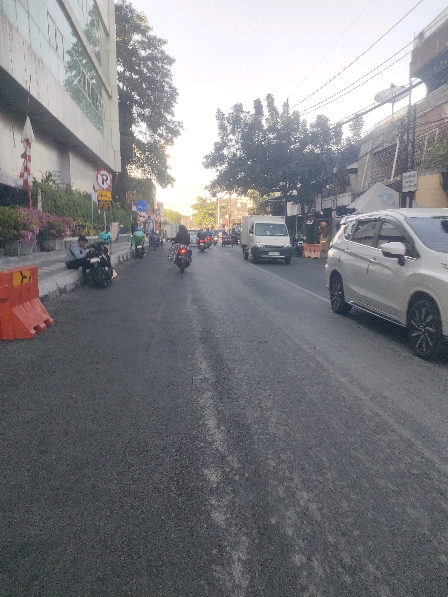 The image depicts a street scene in Surabaya, Indonesia, likely during the daytime as indicated by the natural lighting. The road is paved with asphalt and appears to be a multi-lane street with traffic. On the left side of the street, there is a modern building with large windows and some greenery. In front of the building, there are several motorcycles parked. One person is seated on the curb next to a motorcycle, appearing to be resting or waiting.
Various traffic signs are visible, including a red circular sign with a white "P" inside, indicating parking restrictions. There are also several orange traffic cones placed along the curb.
In the street, traffic consists of several motorcycles and at least one white van. A white SUV is prominent in the foreground, driving on the right side of the road. The sky is overcast or hazy. Electrical wires and poles are visible in the background, suggesting an urban environment. The overall impression is of a busy street in a commercial or residential area of Surabaya.
