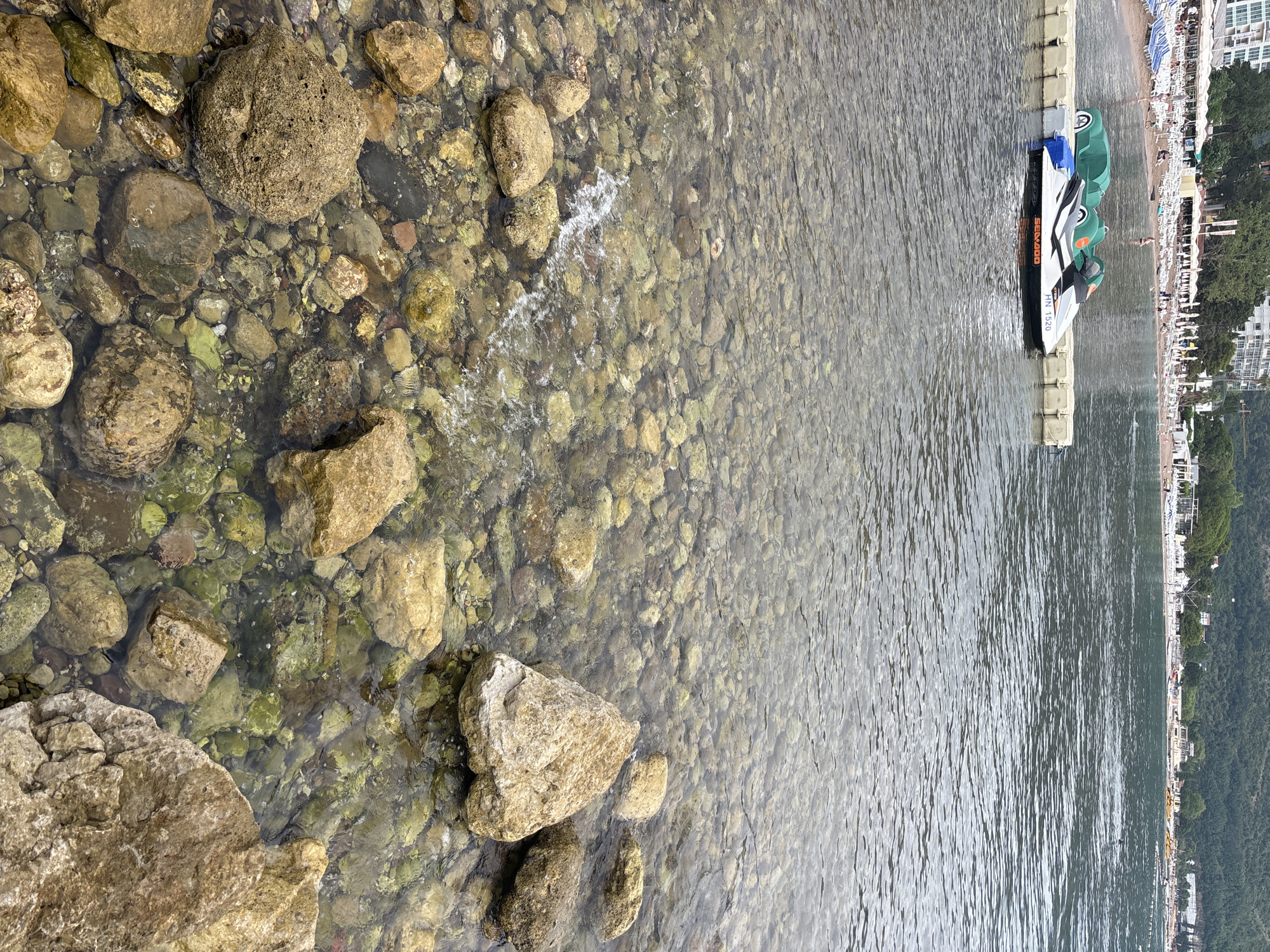 This image depicts a scene along the coast, likely in Budva, Montenegro. The foreground is dominated by shallow, clear water revealing a rocky seabed. Large, weathered stones of various sizes and colors are scattered across the bottom, some partially submerged. The water is calm, with gentle ripples reflecting the overhead light.
To the right, a floating dock is visible, holding several pedal boats. One pedal boat, in white and black with "SEA-DOO" and "NH 1520" printed on it, is prominently featured. The other pedal boats are green. The dock itself appears to be made of modular, light-colored sections.
Beyond the dock and water, a beach area is visible. It is lined with lounge chairs and umbrellas, suggesting a recreational setting. In the background, buildings and lush green hills are present, indicating a coastal town or resort. The sky is not clearly visible, but the lighting suggests it is daytime, possibly late morning or afternoon, under fair weather conditions. The overall atmosphere appears peaceful and relaxed, consistent with a beach vacation destination.