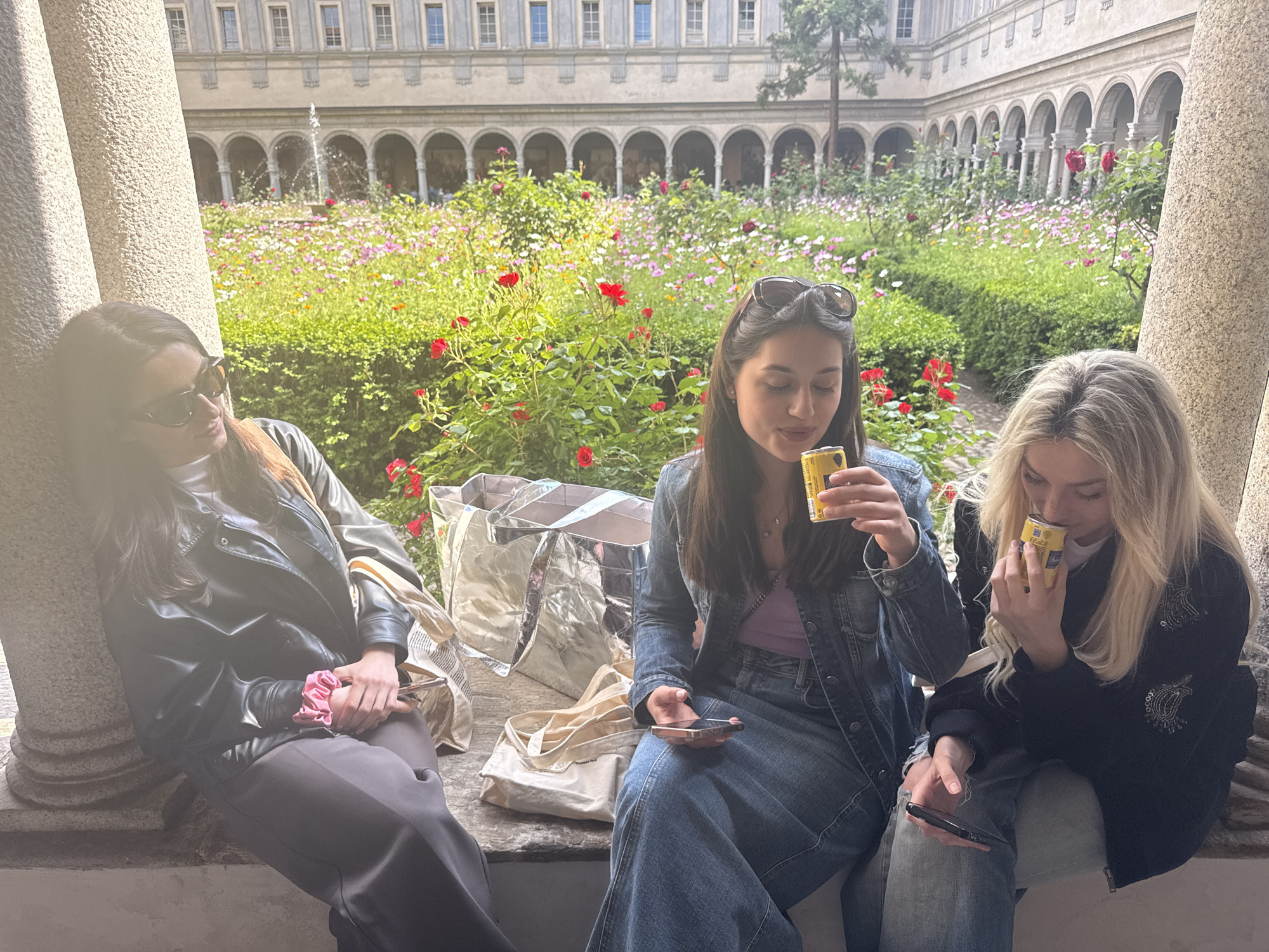 Three young women are casually seated on a stone bench in a sunlit cloister garden in Milan, Italy. The setting features ornate stone columns in the foreground and a historic building with arched walkways and rectangular windows in the background. A lush garden with red roses, various pink and white flowers (possibly cosmos), and green bushes fills the central courtyard, with a small fountain visible in the distance.
The woman on the left, with dark hair and sunglasses, wears a black leather jacket over a white top and dark pants. She leans against a stone column, looking towards the right with a relaxed expression. A pink scrunchie is on her left wrist.
In the center, a woman with dark hair, sunglasses pushed up on her head, is dressed in a denim jacket, a purple top, and wide-leg denim pants. She takes a sip from a yellow Coca-Cola can while holding a smartphone in her left hand, appearing focused on her drink. Behind her is a large, metallic silver tote bag and a beige fabric tote bag with the faint text "Slowdown Studio" visible.
On the right, a blonde woman wears a dark jacket with light embroidered details and light-colored pants. She is also drinking from a yellow Coca-Cola can and holds a smartphone in her left hand, looking down.
The bright daylight suggests a pleasant, warm day. The overall atmosphere is one of relaxed socializing or a leisurely break amidst beautiful architecture and nature.
