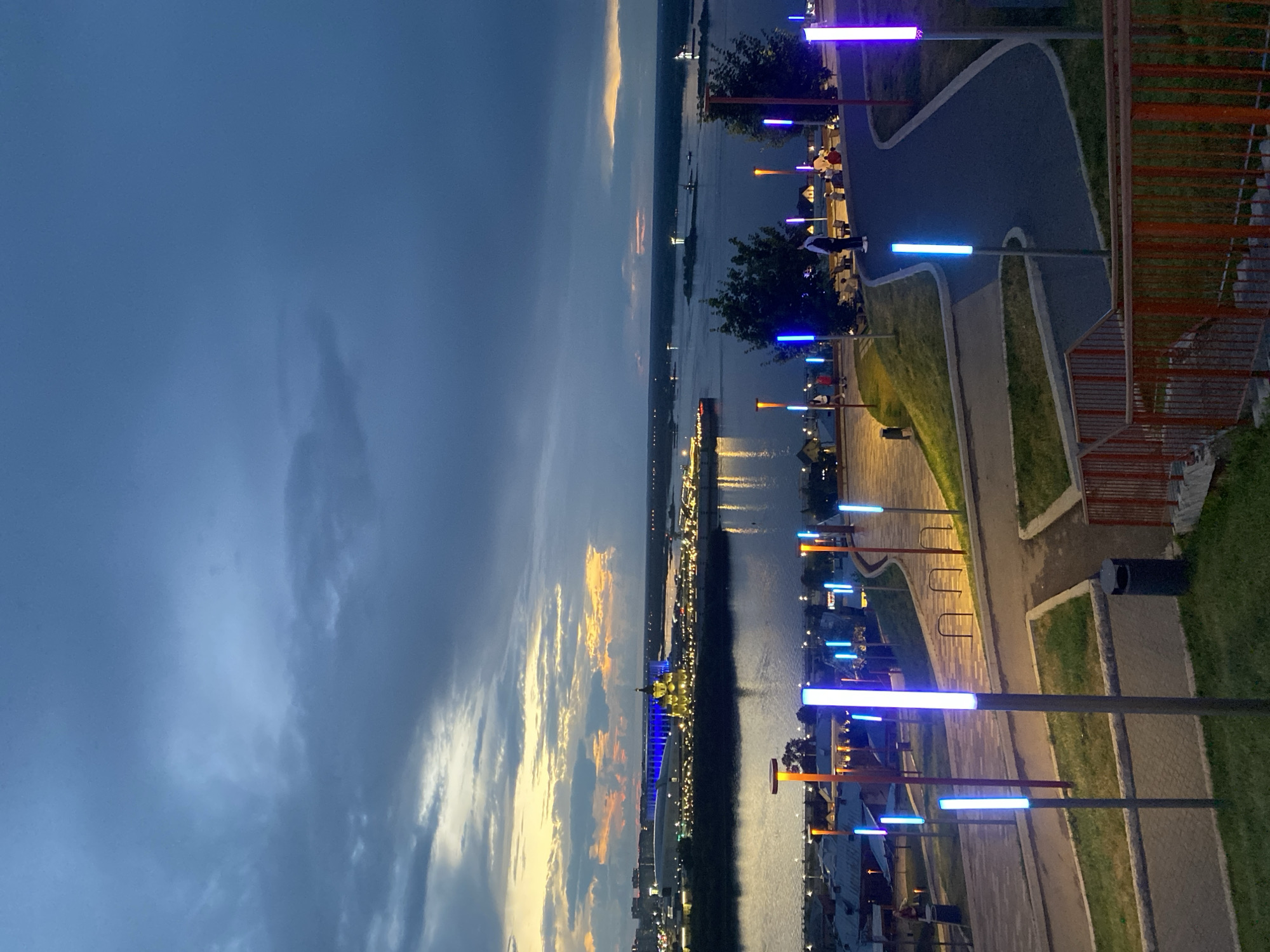The image depicts a view of the Volga River in Nizhny Novgorod, Russia, at dusk. The sky is filled with dramatic, dark clouds illuminated by the fading sunlight on the horizon, creating a gradient of orange and yellow hues. The foreground features a well-lit pathway with numerous modern lamps, some emitting blue light and others orange. People are visible on the pathway, some walking and others sitting on benches, enjoying the view. Across the river, the city skyline is visible, with a prominent illuminated building that appears to be a church or cathedral, and a large stadium with blue lighting. Lights from buildings and a ship on the river also reflect on the water's surface, adding to the atmospheric scene. The overall impression is one of urban twilight, with a blend of natural and artificial illumination.