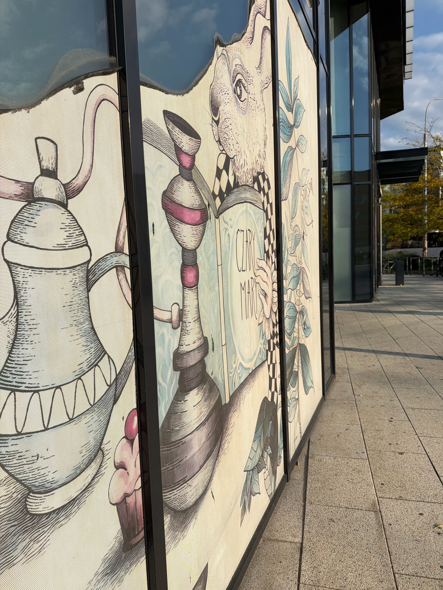 The image shows a mural on the exterior of a building. The mural depicts a whimsical scene with a large, stylized teapot on the left, followed by a detailed illustration of a hookah and then a rabbit reading a book. The book has text that appears to be in Polish, reading "CZARY MARY." The artwork is rendered in a black and white, line-drawing style with subtle pastel accents. Behind the mural, a modern building with large glass windows is visible, reflecting the sky and surrounding trees. The mural is mounted on a perforated screen, allowing some light to pass through. The scene is set outdoors on a paved walkway. This mural is located in Wrocław, Poland, specifically on the exterior of the building housing the University of Wrocław's Faculty of Law, Administration and Economics, situated at 191-192 Plac Grunwaldzki. The time of day appears to be late afternoon, judging by the long shadows cast on the pavement.