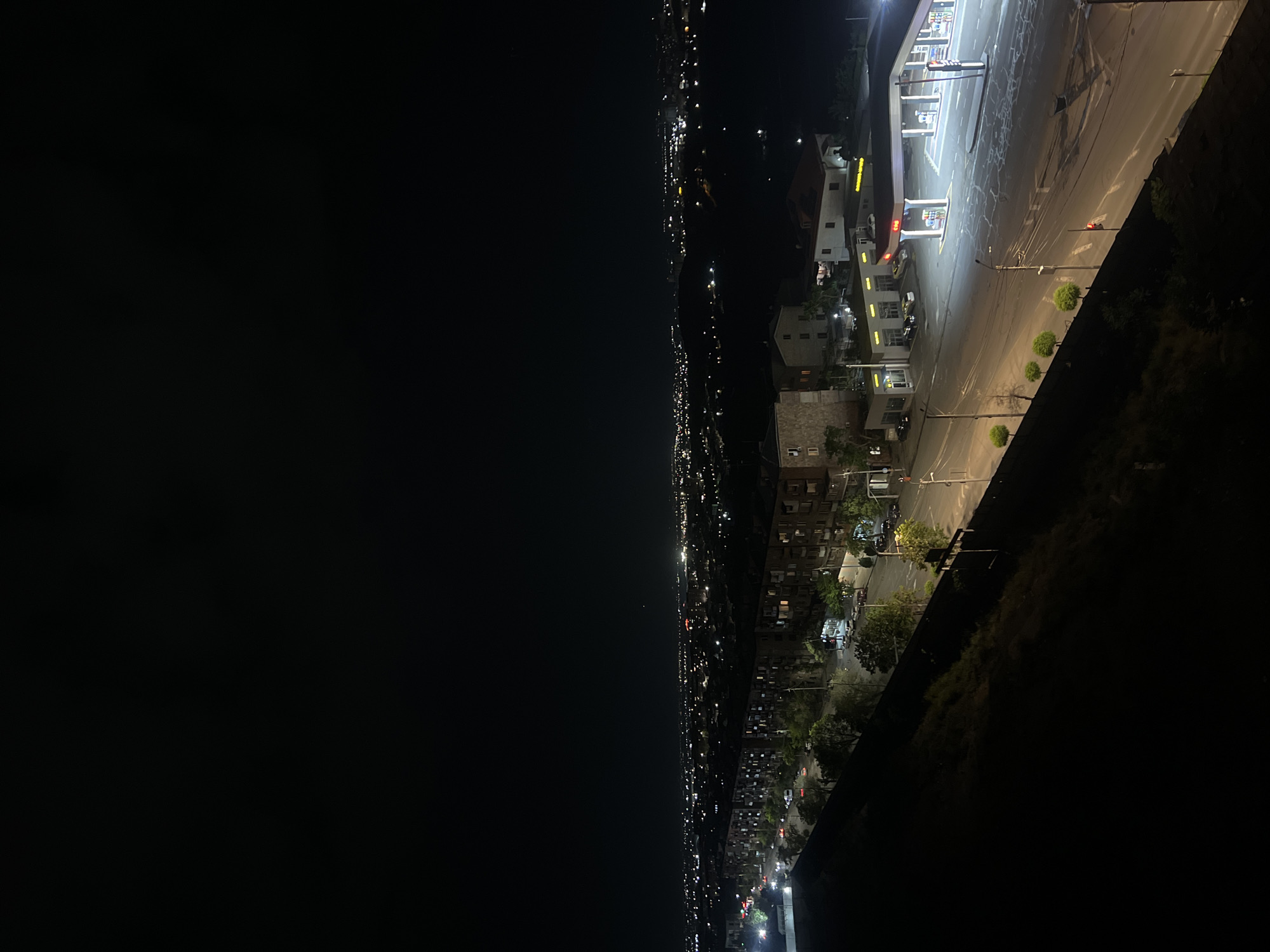 The image shows a night view of Yerevan, Armenia. A brightly lit gas station dominates the upper right. Further into the distance are residential buildings and streets illuminated by lights, extending into the dark horizon on the left. The foreground includes a wall or embankment with vegetation.