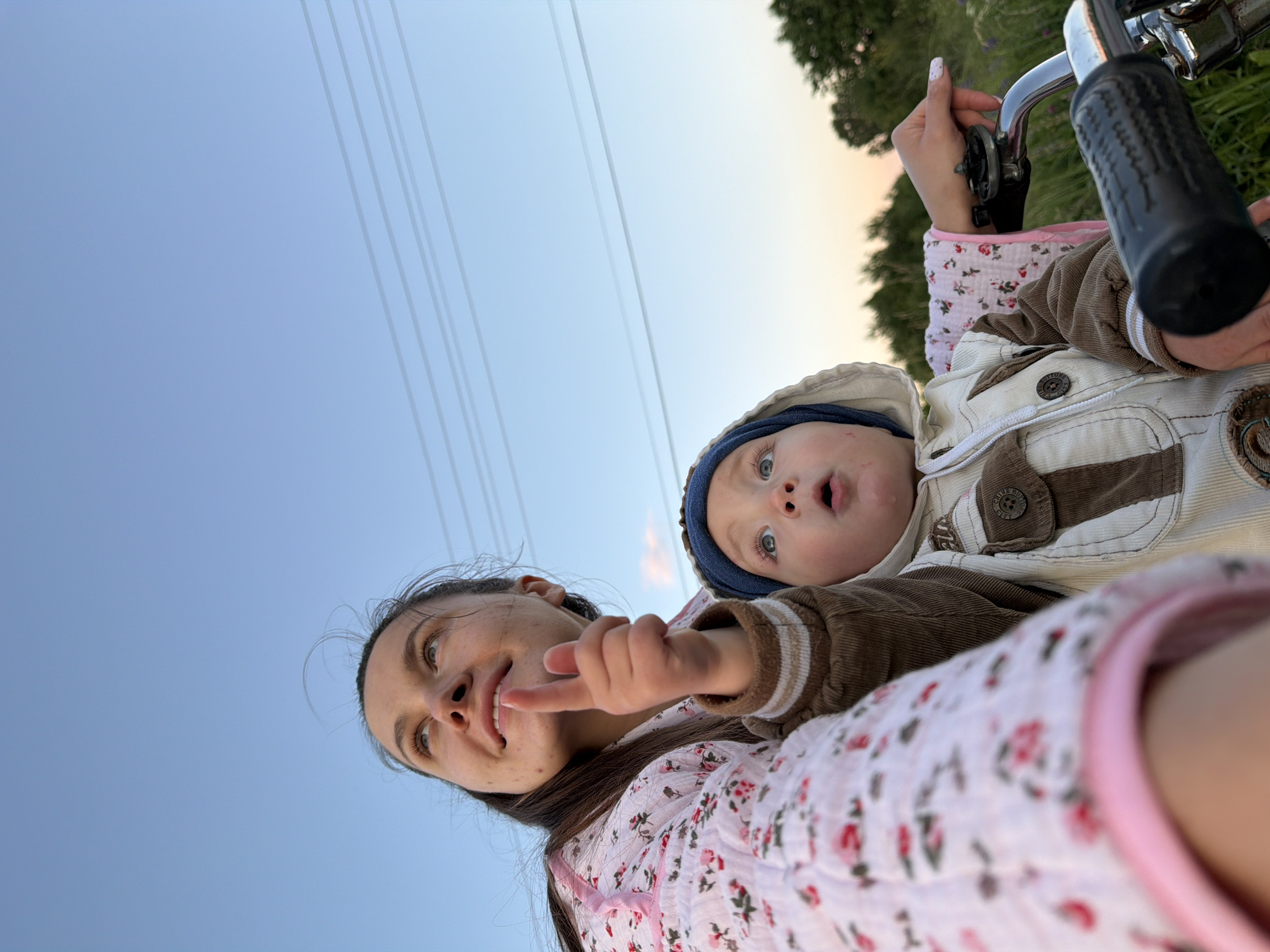 A young woman and a baby are sitting on a bicycle on a dirt road in the village of Krasny Mayak, Russia. The woman is looking up at the sky, pointing with her index finger. The baby is looking up at the sky with its mouth open. The background is a field of grass and trees. The sky is blue with some clouds. The woman is wearing a pink and white floral dress. The baby is wearing a brown and white jacket with a blue hood. The bicycle has a black handle and a silver frame.
