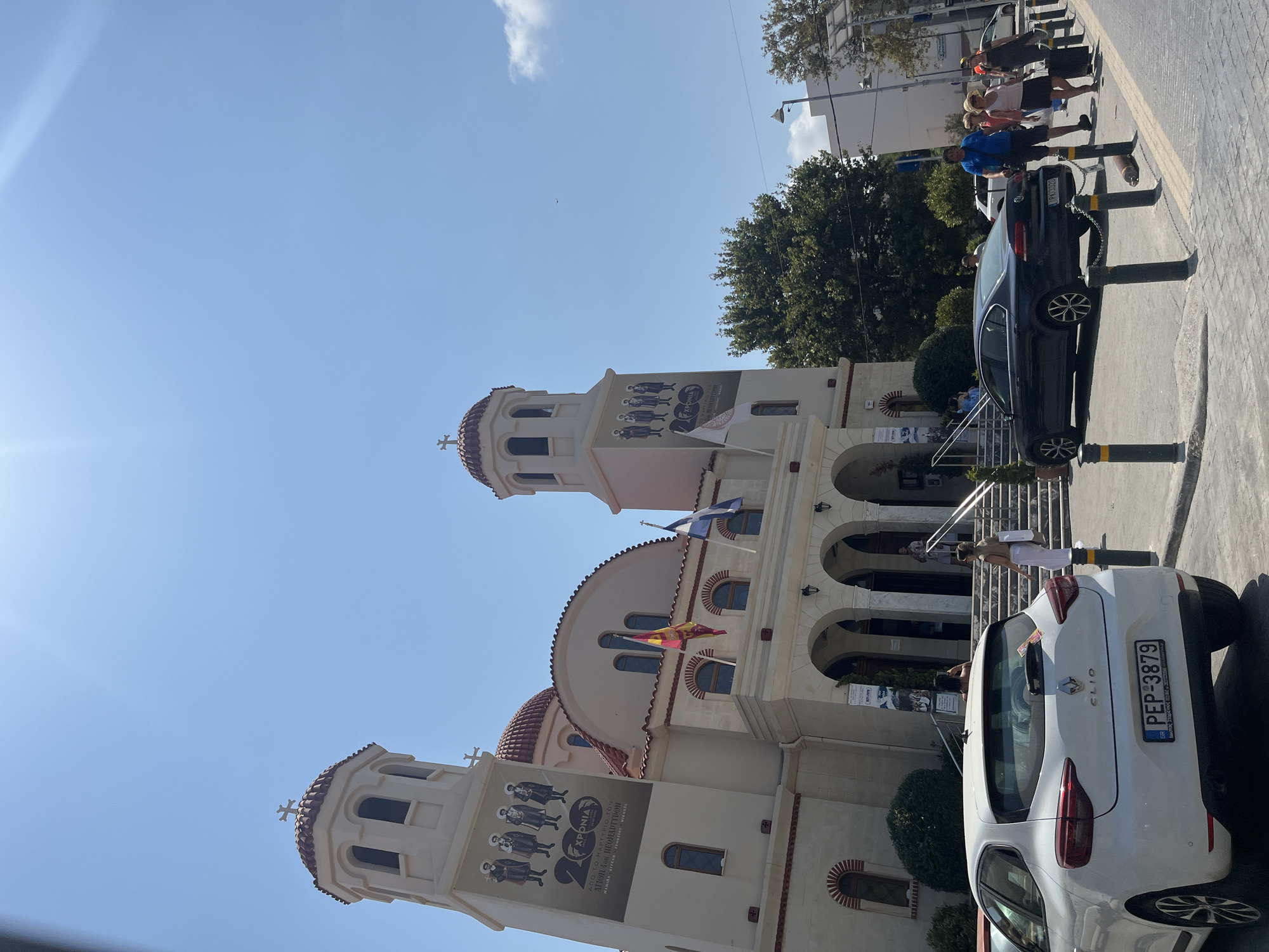 A bright, sunny day with a clear blue sky is depicted, with a slight lens flare in the upper right corner. The foreground features the rear of a white Renault Clio, license plate PEP-3879. To its right, a dark sedan is parked. Behind the cars, the facade of a large, light-colored church with red-tiled domes and bell towers is prominent. A banner with figures and Greek text is visible on the left bell tower. Two flags, one red and yellow and the other blue and white, fly near the entrance arches. Steps lead up to the church entrance, where several people are visible. To the right of the church, trees and a street lamp are present, with a white building in the background. The street in front of the church is paved with light-colored cobblestones and bordered by a yellow line. Several people are walking on the sidewalk to the right of the parked cars. The scene is likely in Rethymno, Greece, given the architectural style and flags.
