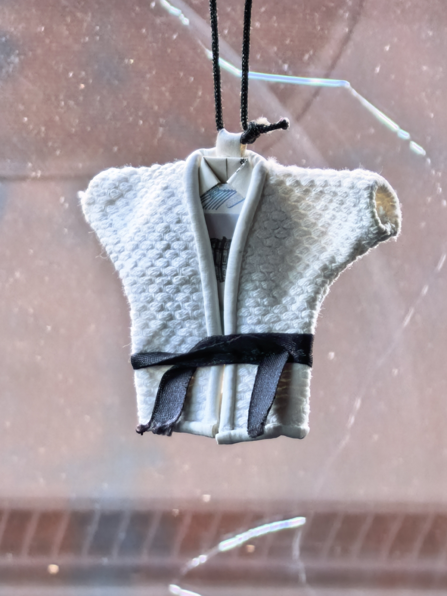 A miniature white gi, a type of martial arts uniform, is hanging by a black cord. The gi is detailed with a textured fabric and a black belt tied around the waist. The background is blurred and appears to be a window with light reflections, suggesting it's indoors or near a window. There are no people visible in the image. The scene is static and the gi is the sole focus of the photograph. No specific location in the city of Sterlitamak, Russia can be determined from the image.