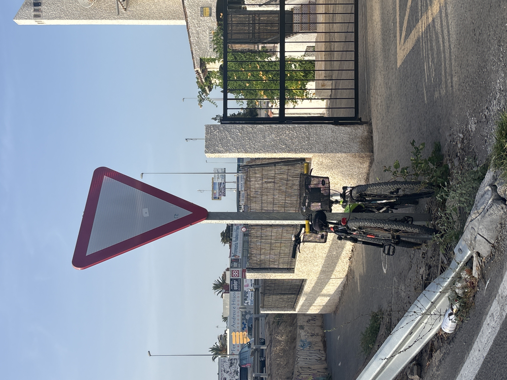 A street scene in Orihuela, Spain, features a prominent inverted red-bordered triangle yield sign. Two bicycles are parked near the sign. In the background, a building with a textured white facade and a tall, slender tower is visible, with a dark metal gate in front of it. Adjacent to the building is a fence made of bamboo panels. Further in the distance, a highway with vehicles and commercial signage can be seen, including what appears to be a "Ferreteria Andreu e Hijos" sign. The ground is paved, with some debris and overgrown vegetation along the edges. The sky is clear and blue, indicating daytime.