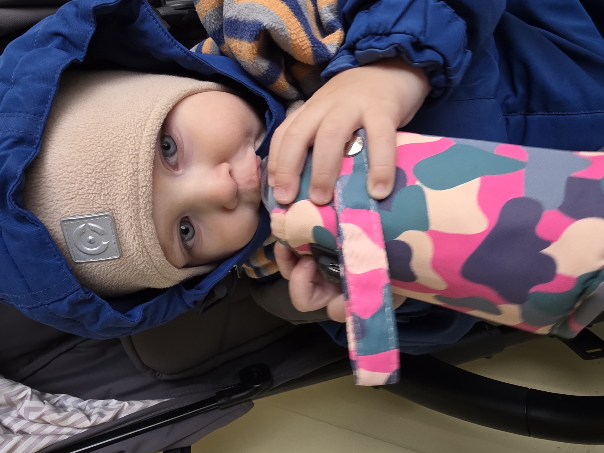 An adorable baby or toddler is captured in a close-up, nestled warmly within a stroller. The child wears a royal blue hooded jacket and a soft, textured beige hat that covers their ears. Piercing blue eyes gaze directly towards the viewer with a calm, curious expression. A small, square silver label, featuring a circular logo with a stylized, intertwined 'C' or similar design, is visible on the front of the hat.
The baby's left hand is seen gripping an object or accessory adorned with a vibrant camouflage pattern in shades of pink, teal, light blue, grey, and beige. A striped, warm-looking blanket or scarf in orange, blue, and grey is also visible near the baby's head. The interior of the stroller, including grey fabric and a patterned blanket with white and light pink stripes, fills the lower left of the frame.
The clothing suggests cool or cold weather, suitable for an outing in Chaikovsky, Russia. The scene appears to be taking place during the daytime, likely outdoors or in a well-lit indoor space, conveying a moment of cozy comfort.
