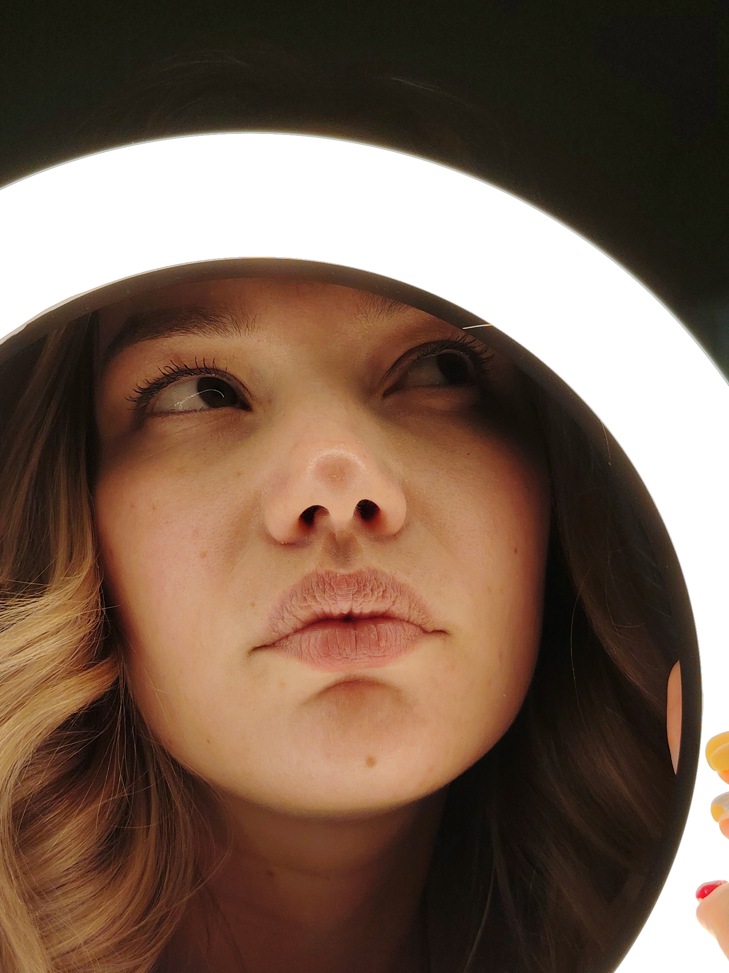A close-up shot features a woman's face illuminated by a ring light. She looks upward and to the right, with her eyes slightly wide and her lips closed. Her hair is visible on the sides, showing a blonde color with soft waves. The environment outside the ring light is dark. The image was taken in Марковский, Russia.