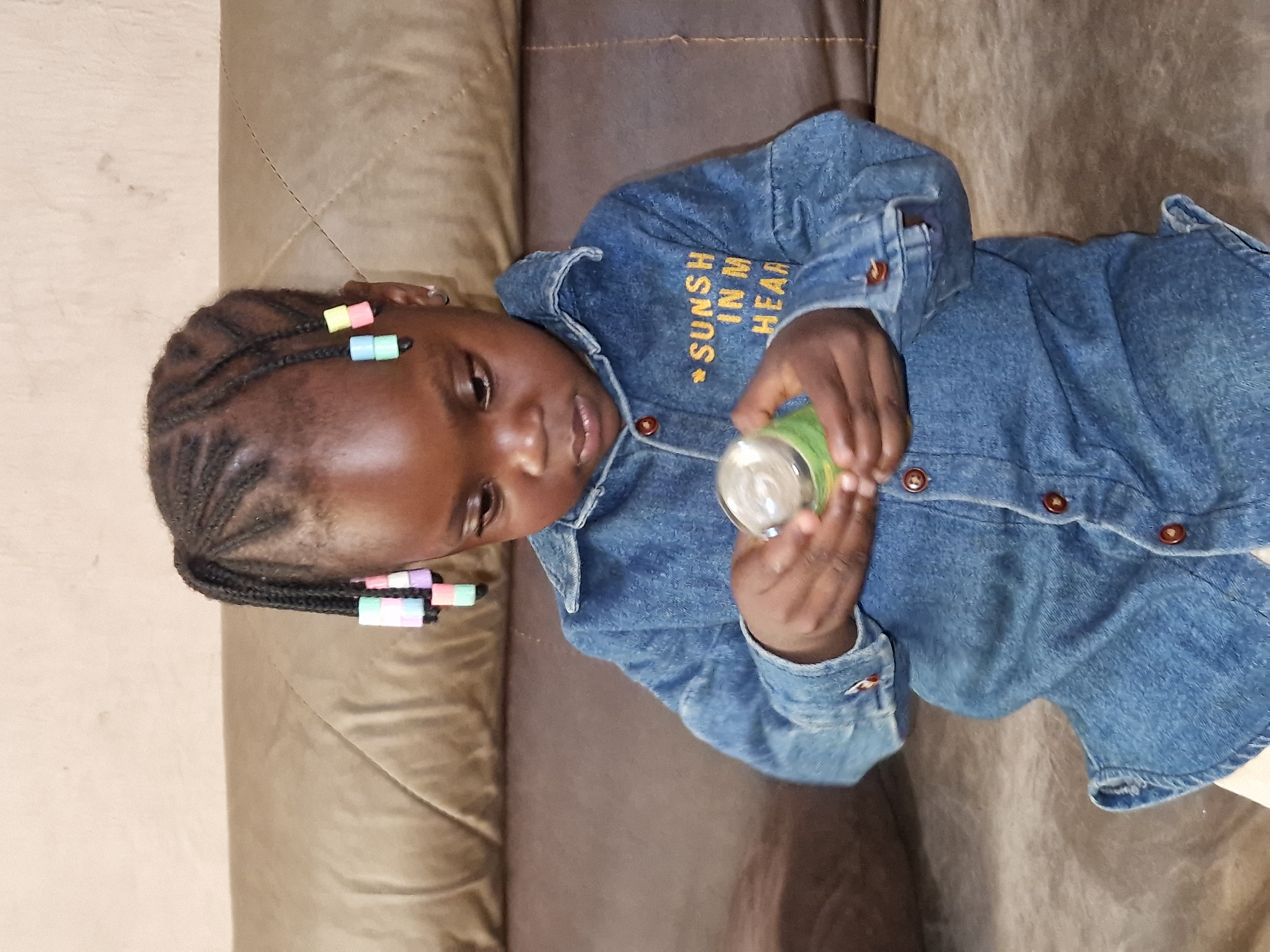 A young Black girl with braided hair adorned with colorful beads sits on a brown couch. She is wearing a blue denim shirt with buttons and a yellow text design that reads "SUNSH IN M HEAR". The girl is holding a small green cylindrical object in her hands and appears to be examining it. The background consists of a textured wall and the couch. The setting is an indoor residential environment. The presence of the couch and the general room decor suggest a home environment in the city of Jos, Nigeria.