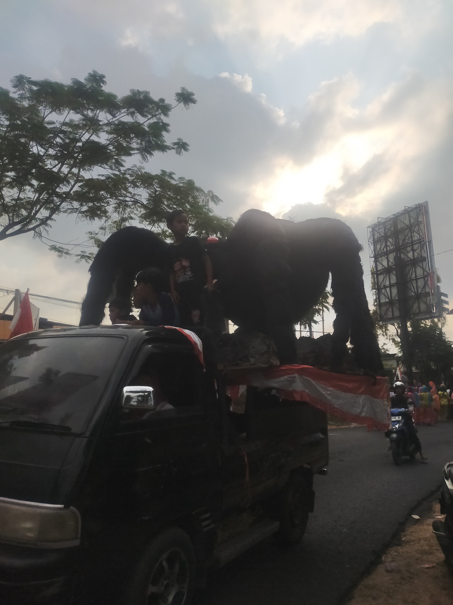 In Tuwel, Indonesia, a black pickup truck is parked on a street, possibly during a parade or festival. The truck is carrying two large, dark-colored, gorilla-shaped figures. Two young boys are standing on the back of the truck, one behind the other, with the boy in front wearing a dark t-shirt with graphics. A third child, also wearing a blue shirt, is partially visible behind the first boy. The truck's bed is decorated with a red and white striped fabric, resembling the Indonesian flag. In the background, trees, a large billboard structure, and a cloudy sky with the sun peeking through are visible. A motorcyclist wearing a helmet rides on the street, and other people in colorful clothing can be seen further down the road. The overall scene suggests a festive or cultural event taking place outdoors.