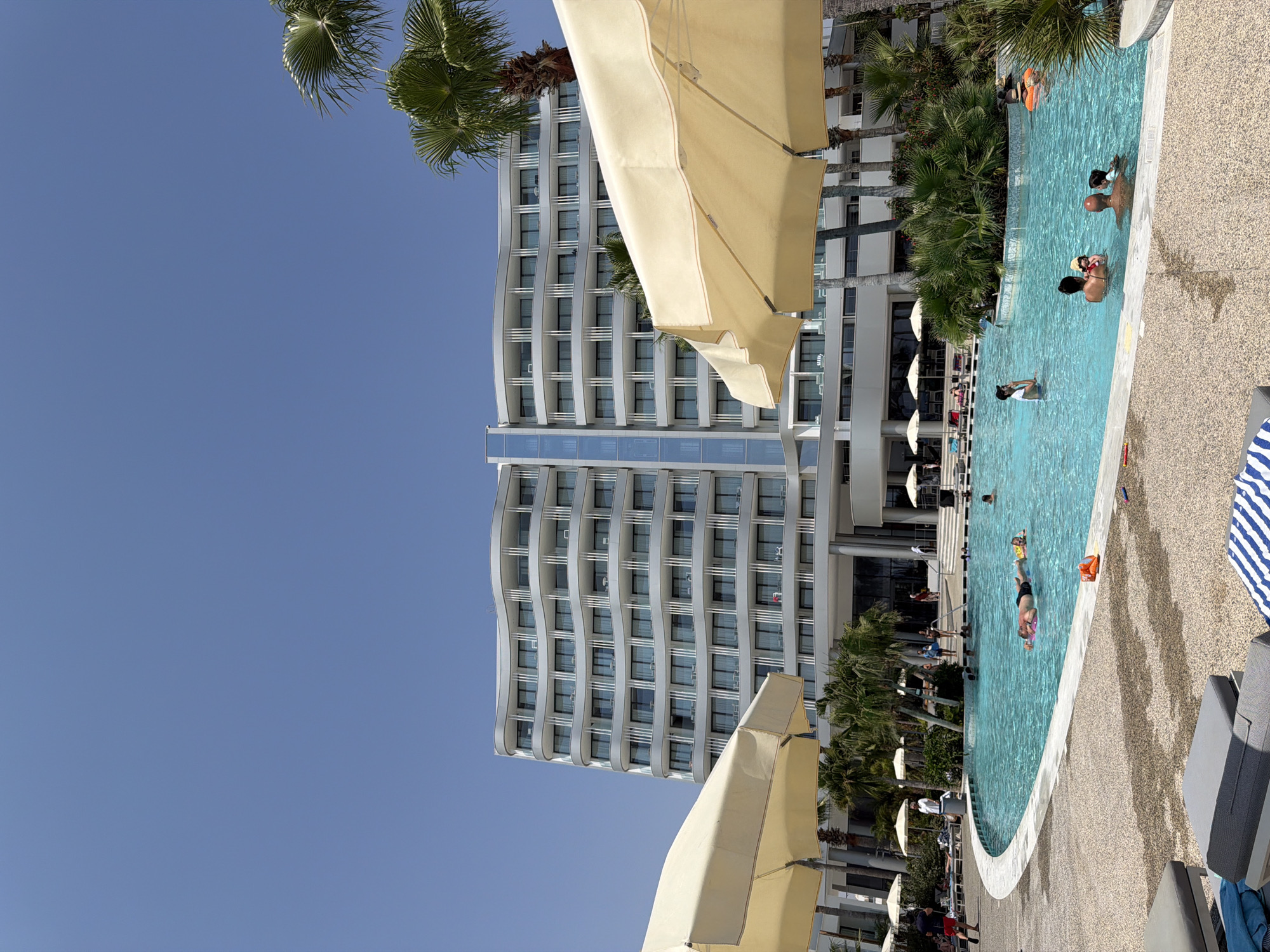 A wide shot captures a sunny day at a resort pool in Oroklini, Cyprus. A modern, multi-story hotel with a curved facade dominates the background under a clear blue sky. In the foreground, a large, irregularly shaped swimming pool is filled with people enjoying the water. Palm trees are scattered around the pool area, and large, cream-colored umbrellas provide shade over lounge chairs on the paved deck. Several individuals are visible swimming, floating on inflatable toys, or standing in the shallow end of the pool. Some people are relaxing on lounge chairs on the pool deck, and a few others are walking near the pool. The overall scene depicts a leisurely resort atmosphere on a bright, warm day.