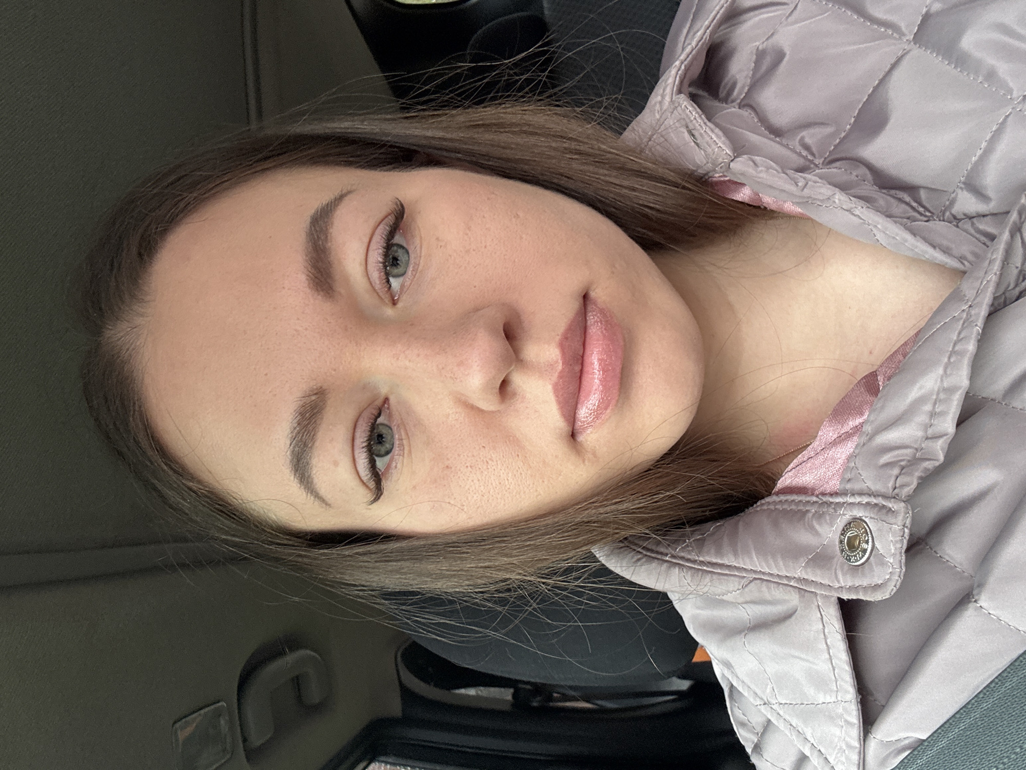 A young woman with light brown hair and light eyes is facing the camera. She has fair skin with freckles visible on her face. She is wearing a light purple quilted jacket with a pink inner lining and a silver button closure. The background is dark and suggests that she may be inside a vehicle. There is a car door handle visible behind her head. The location is Ферма, Russia.