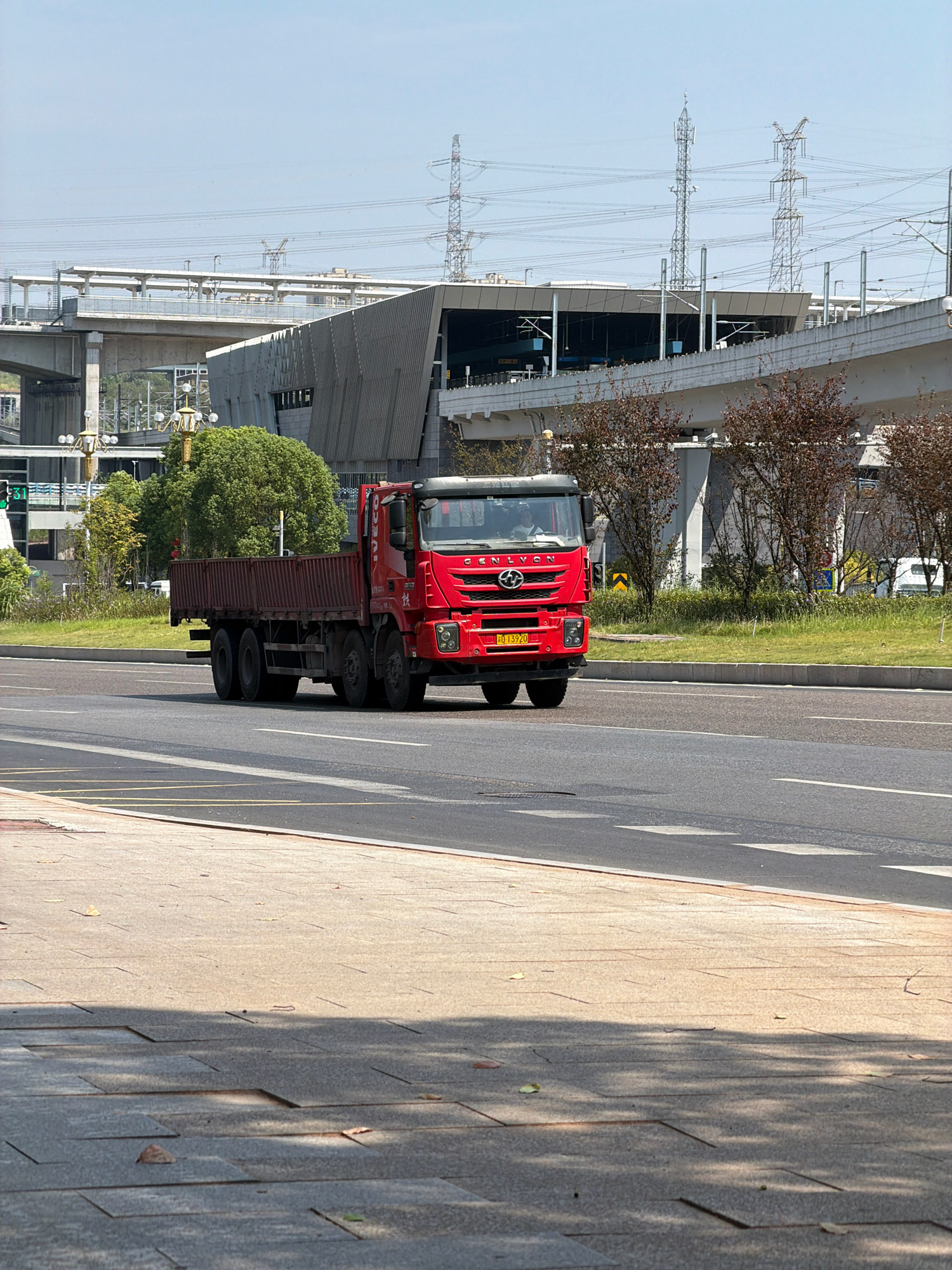 A red flatbed truck with a dark brown cargo bed is driving on a road in Jiangjin, China. The truck has six wheels, with a visible axle on the rear of the vehicle. The truck's windshield shows a driver wearing a white shirt. The truck is branded with the logo "GENLYON" on the front. The license plate of the truck is yellow with black characters, displaying "0 13920". The truck is on a multi-lane road, with other vehicles visible in the background. A modern building with a distinctive angular facade is situated behind the truck, with elevated infrastructure, possibly a railway or highway overpass, extending from it. There are power lines and transmission towers in the background. The scene appears to be an urban environment with a mix of infrastructure and greenery, suggesting a well-developed area. The foreground shows a pedestrian walkway paved with rectangular stones, with shadows cast by trees. The sky is clear and blue.
