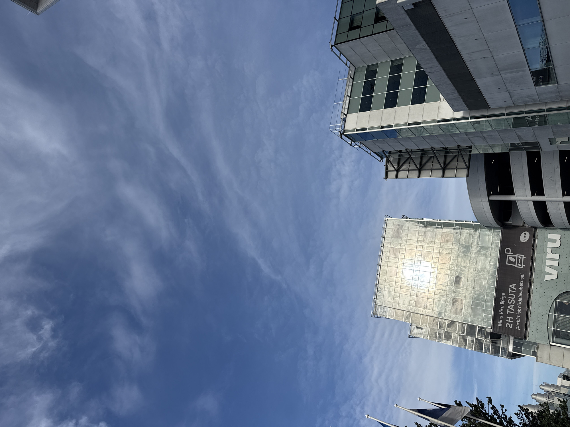 The image shows a low-angle view of modern buildings against a partly cloudy blue sky. On the left, there are flags and trees. One building, on the left, has a reflective glass facade, and a banner below it reads "VIRU" and advertises "2H TASUTA parkimist nädalavahetusel" (2 hours free parking on weekends). Another building, to the right, is made of concrete and glass. A circular parking garage structure is visible between the two buildings. The overall scene depicts an urban environment with contemporary architecture.