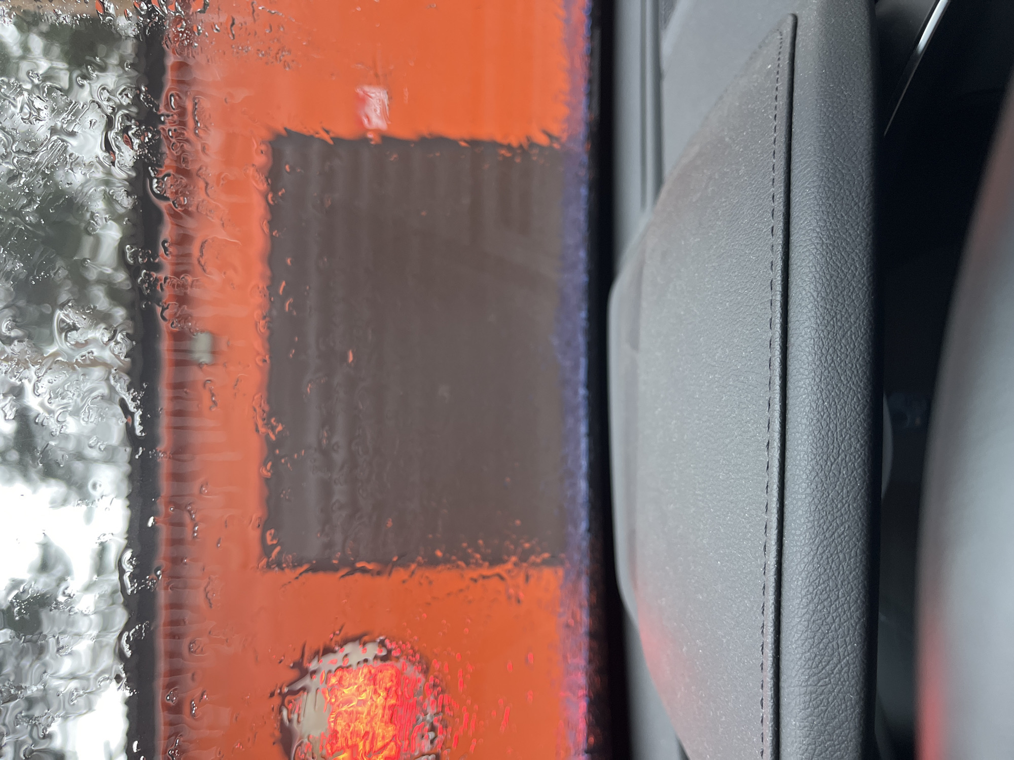 The image shows a view from inside a car, looking out through the driver's side window. The window is covered in raindrops, obscuring the view. Through the wet window, there is a blurred view of what appears to be an orange and grey structure, possibly a building. The interior of the car is visible on the right side of the image, showing a section of the car door. The location in the city of Chaikovsky, Russia, is not identifiable from the image.