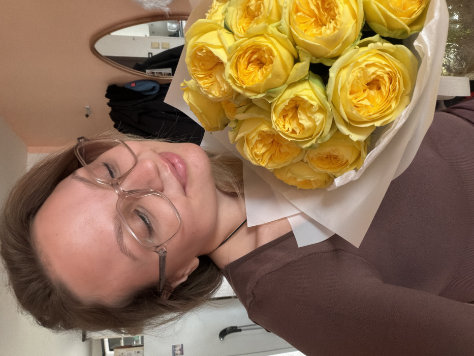 A female individual with blonde hair and clear-framed eyeglasses is depicted indoors in a residential setting in Tel Aviv-Yafo, Israel. She wears a dark brown top and a thin dark necklace. Her eyes are closed, and her lips are slightly parted in an expression suggesting sensory engagement. She holds a bouquet of approximately a dozen vibrant yellow roses, wrapped in white paper, positioned near her face. The background on the left features a white refrigerator door. The background on the right shows a peach-colored wall with a narrow, arch-topped mirror reflecting part of a kitchen area, including a metallic kettle. Dark clothing items hang below the mirror.