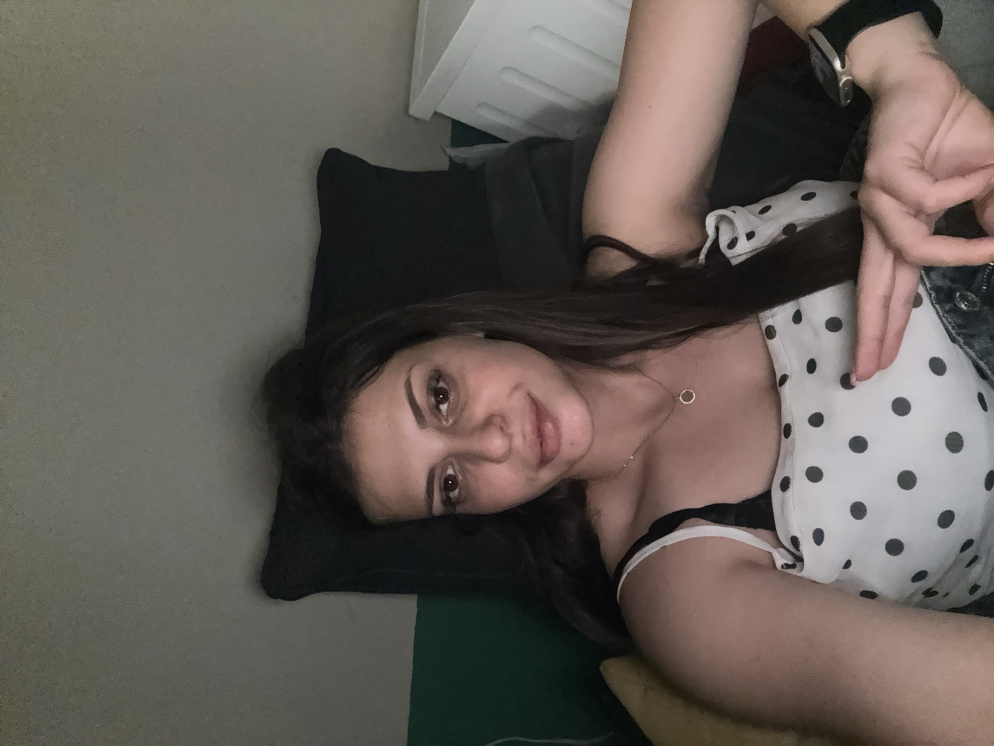 The image is a selfie taken by a woman with long, dark hair and brown eyes. She is wearing a white and black polka dot top with thin straps and a delicate necklace with a circular pendant. She has a smartwatch on her left wrist. Her left arm is resting on her lap, and her right hand is positioned near her chest, with her index finger and thumb forming a circle, while her other fingers are slightly extended. The woman appears to be indoors, possibly reclining on a sofa or bed, with dark pillows visible behind her. To her right, a white storage unit with a textured surface is partially visible. The lighting is dim, suggesting an evening or indoor setting. The image is cropped to focus on the woman's upper body and face. The scene does not provide any visual clues to its specific location within the city of Plovdiv, Bulgaria.