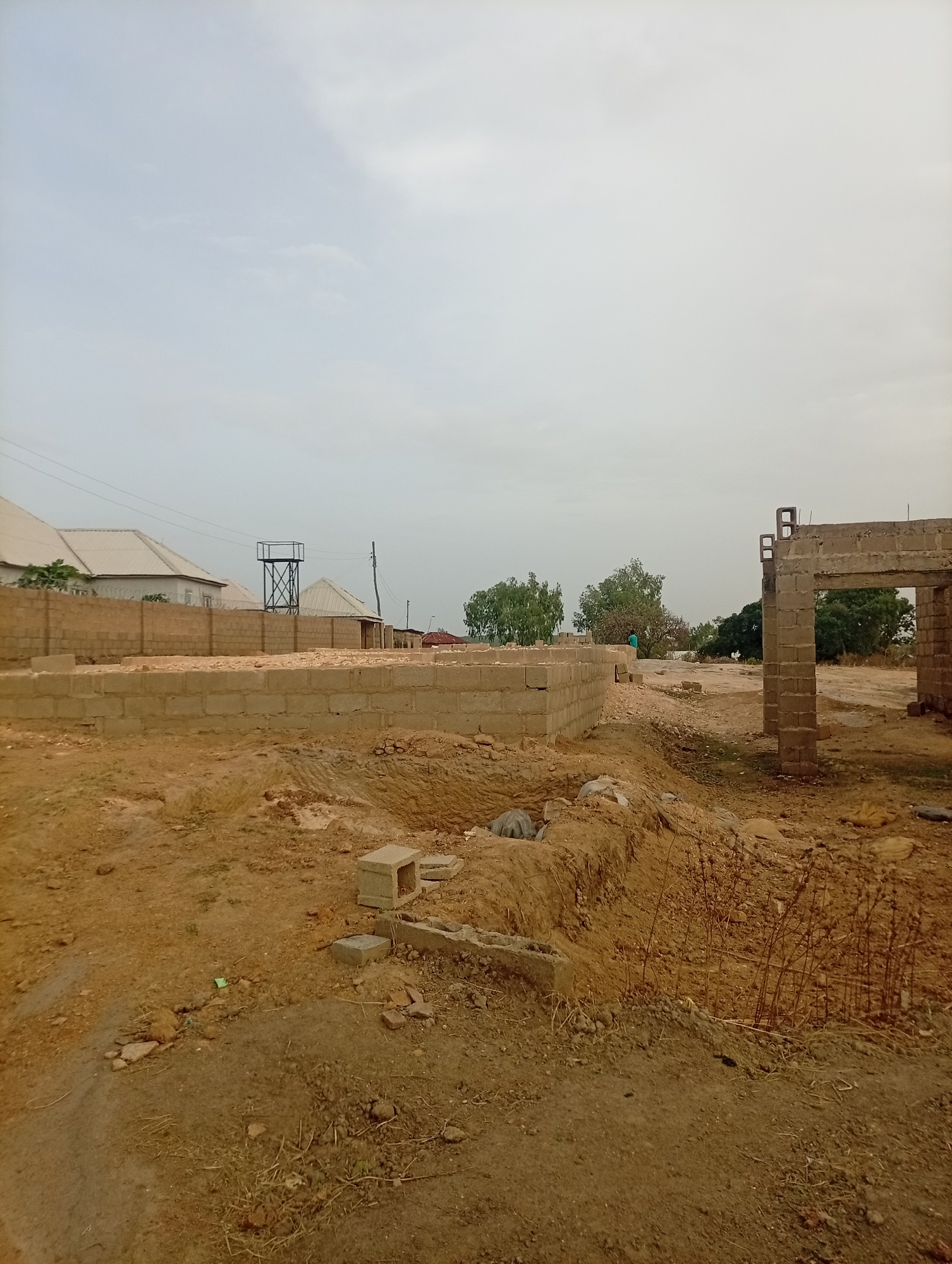 The scene depicts a construction site in Jos, Nigeria. In the foreground, there is a mound of brown soil with sparse vegetation and scattered building materials, including concrete blocks. The partially constructed foundation of a building extends into the background, consisting of multiple courses of cinder blocks. Beyond the foundation, a water tower and several buildings are visible in the distance, set against a cloudy sky. A partially constructed, block pillar structure stands to the right of the frame. A person wearing a green shirt is observed in the mid-ground.