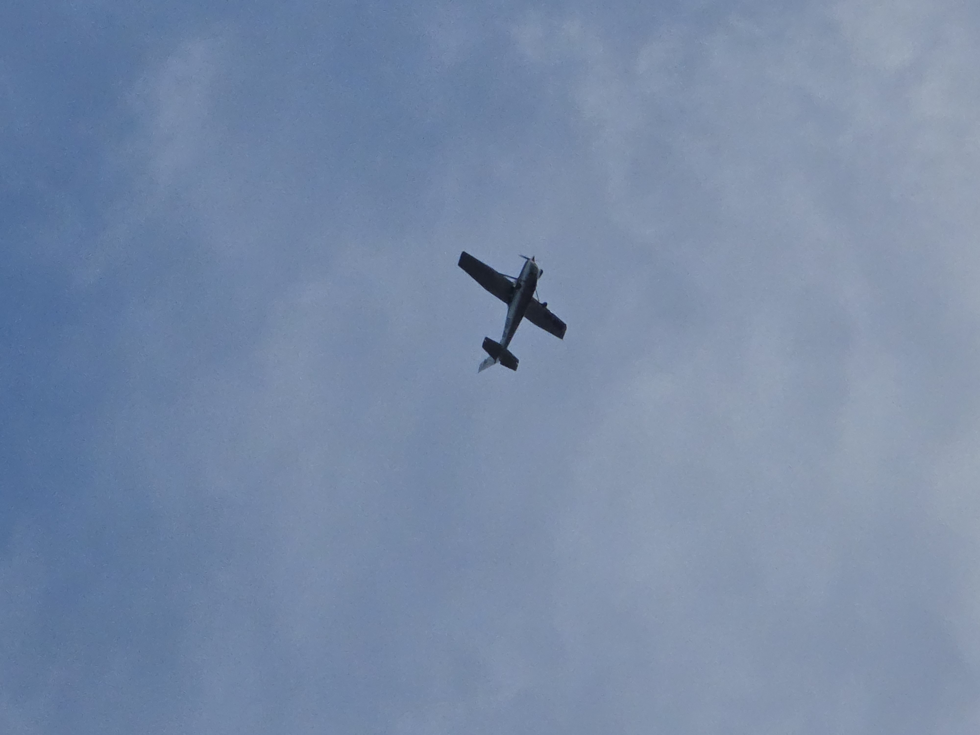 A small, single-engine airplane is captured in flight against a hazy blue sky. The aircraft, appearing dark in silhouette, is viewed from below and slightly to the side, showing its wings, fuselage, and tail. Its propeller is visible at the front, suggesting it is powered. The plane is positioned in the upper right quadrant of the image, with the vast expanse of the sky filling the rest of the frame. There is no visible indication of its location in Warsaw, Poland, or any specific environmental context beyond the sky itself.