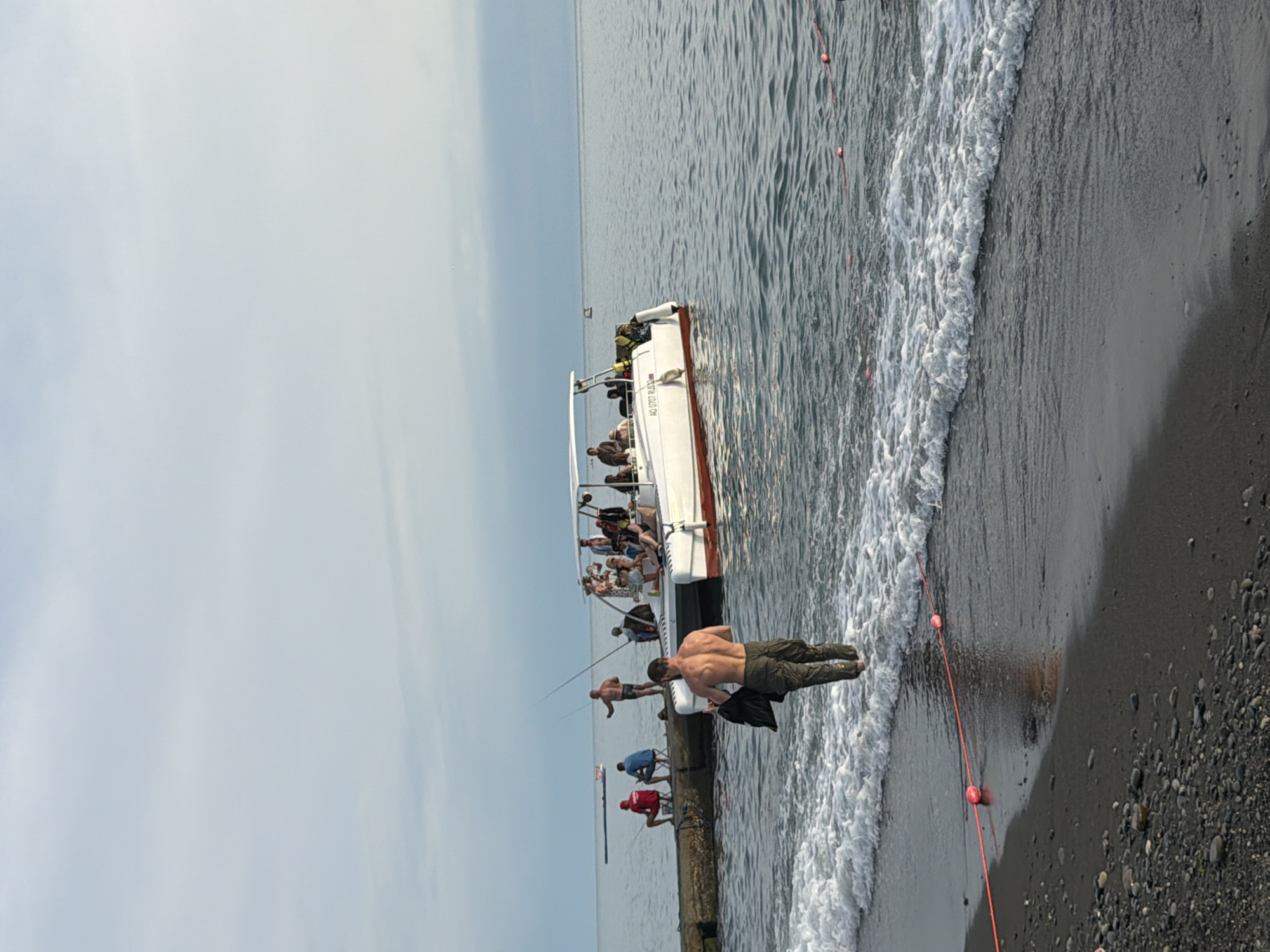 A wide shot captures a scene at the Black Sea coast in Sochi, Russia, near the Adler pier. In the foreground, a man, shirtless and wearing green cargo pants, walks out of the water towards the pebbled beach, carrying a dark bag. Gentle waves crash onto the shore, with a red rope and buoys marking an area in the water.
Further out, a concrete pier extends into the sea, populated by several individuals. Some are fishing with rods, while others are seated or standing. A white catamaran-like boat with a canopy is docked near the end of the pier, carrying a group of passengers who appear to be enjoying a boat trip. The sky is overcast with a pale blue hue, and the sea is calm. The overall atmosphere suggests a leisurely day by the coast, with various recreational activities taking place simultaneously.