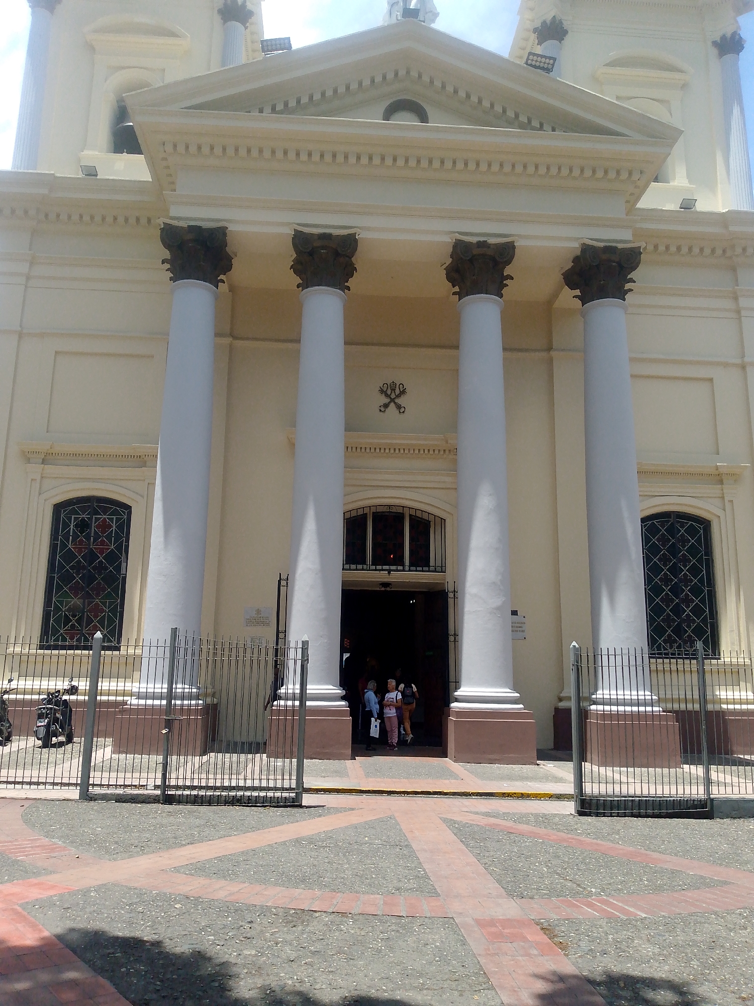 The image shows the front facade of the Caracas Cathedral in Venezuela. The cathedral has a pale yellow facade with four large, white columns supporting a triangular pediment. The columns have dark capitals. There are two arched windows with decorative grilles visible on either side of the entrance. Above the entrance, there is a symbol featuring crossed keys. A wrought iron fence stands in front of the cathedral, partially obscuring the entrance. Several people are entering and exiting the cathedral. The foreground shows a stone-paved area with red brick accents. A motorcycle is parked behind the fence to the left of the entrance. The sky is visible at the top of the frame.