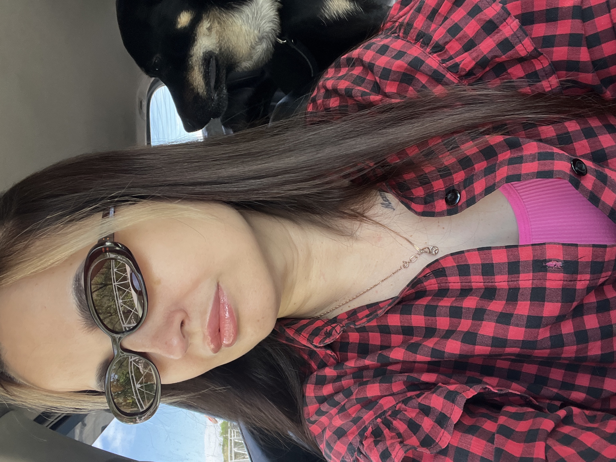 The image is a selfie taken by a young woman inside a vehicle, with a dog partially visible behind her. The photo is rotated 90 degrees counter-clockwise.
The primary subject is a young woman with long, straight, dark brown hair, featuring blonde highlights framing her face. She is wearing dark, oval-shaped sunglasses that reflect outdoor scenery, including trees and what appears to be a white structure against a bright blue sky. Her lips are adorned with glossy, light pink lip gloss. She is dressed in a red and black buffalo plaid button-up shirt, worn open over a ribbed, hot pink tank top. A delicate, gold-toned necklace with a small charm hangs around her neck, and a small, dark tattoo (text or symbol) is visible on her upper chest/collarbone area.
Behind and slightly above the woman, the head and upper back of a dog are visible. The dog has black fur with tan markings, consistent with breeds like a German Shepherd.
The setting is an interior of a vehicle, suggested by the background elements and the close-up nature of the selfie. The bright reflections in the sunglasses indicate it is daytime with clear weather. The woman has a neutral and calm expression. No clearly legible text is visible other than the faint tattoo and a small, unidentifiable logo on her shirt.