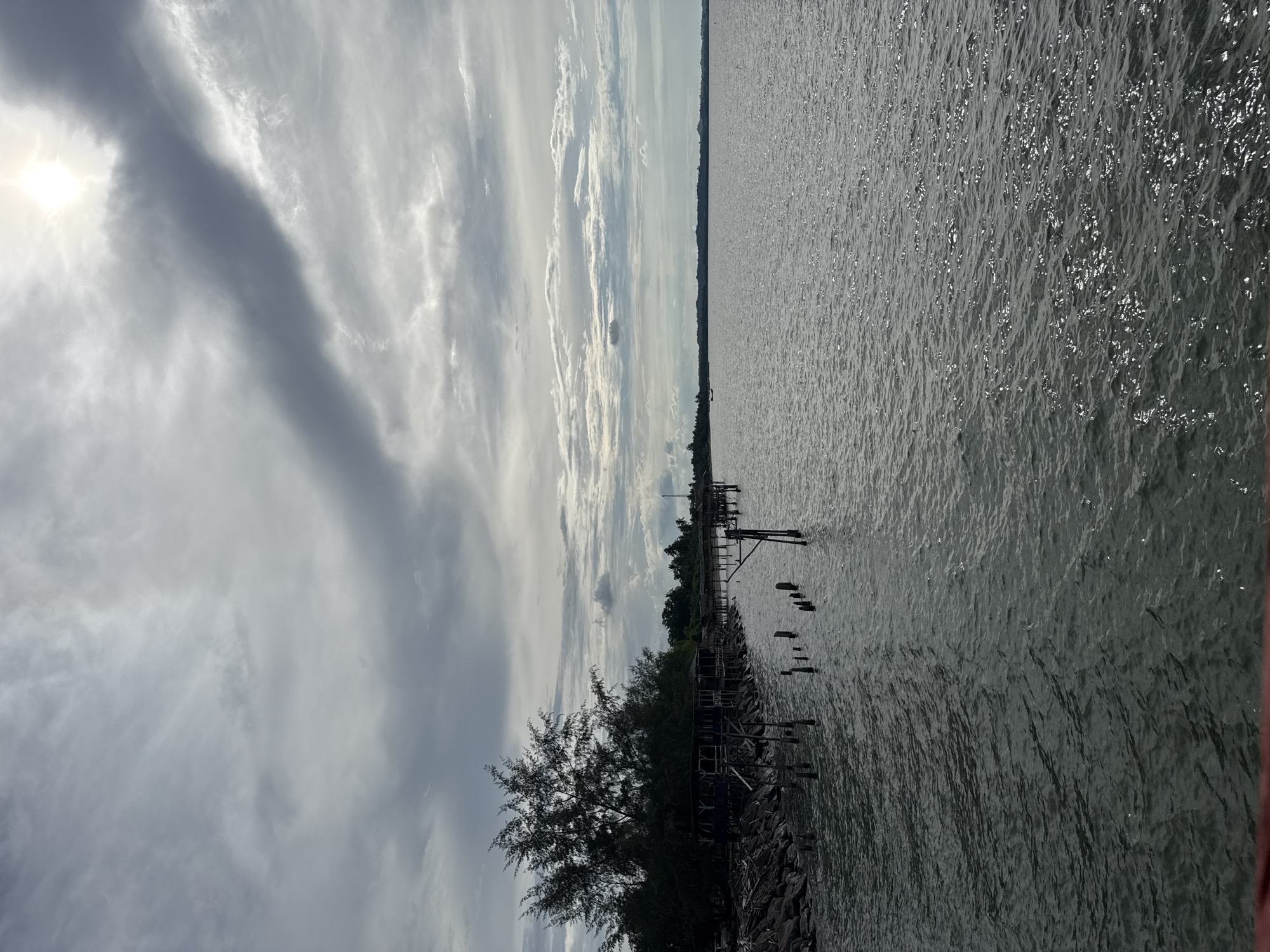 The image shows a body of water in Lancok, Indonesia, with a cloudy sky overhead. The water is rippled, reflecting light. A pier or series of structures extends into the water, with some posts and platforms visible. Trees and vegetation line the shore. The scene appears to be taken during the day.