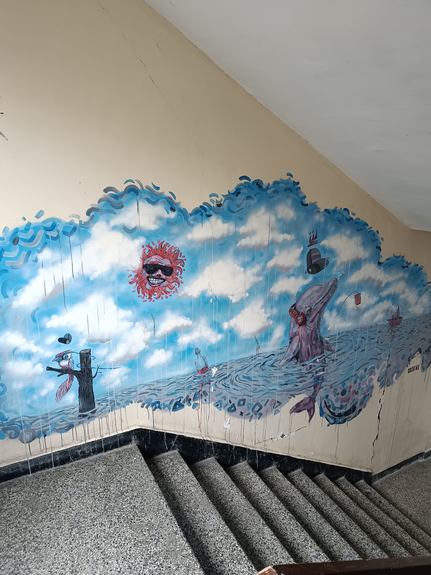 The image depicts a mural on a staircase wall. The mural features an ocean scene with a cartoonish sun wearing sunglasses, a bird on a tree stump with a hat, a dolphin wearing headphones and a crown hat, a bottle, a ship, and rain lines. The wall is cream-colored with cracks. The staircase is made of a speckled gray material with a black border. The location is Sofia, Bulgaria.