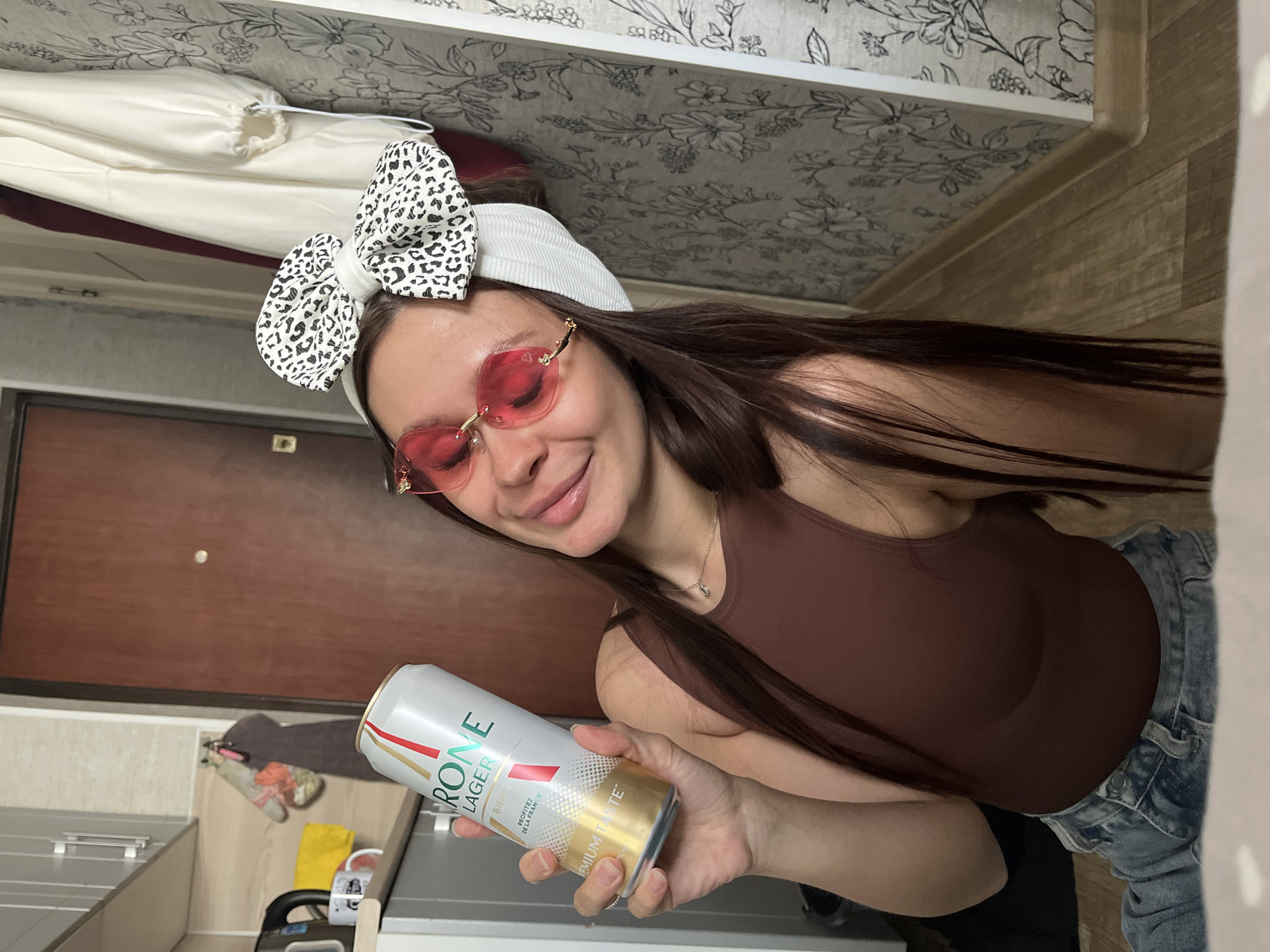 A young woman with long, dark hair, wearing pink-tinted heart-shaped sunglasses and a white headband with a black and white leopard-print bow, smiles at the camera. She is holding a can of "Kronenbourg 1664 Lager" beer in her right hand. She is wearing a brown tank top and blue jeans. The background shows an indoor setting with wooden flooring and a brown door. There are also glimpses of a kitchen counter and cabinets. The lighting suggests it is daytime. The woman appears happy and relaxed. Text visible on the can includes "KRONENbourg," "LAGER," "BIERE," "PROTÉGEZ DE LA FRANCE," and "PREMIUM TASTE."