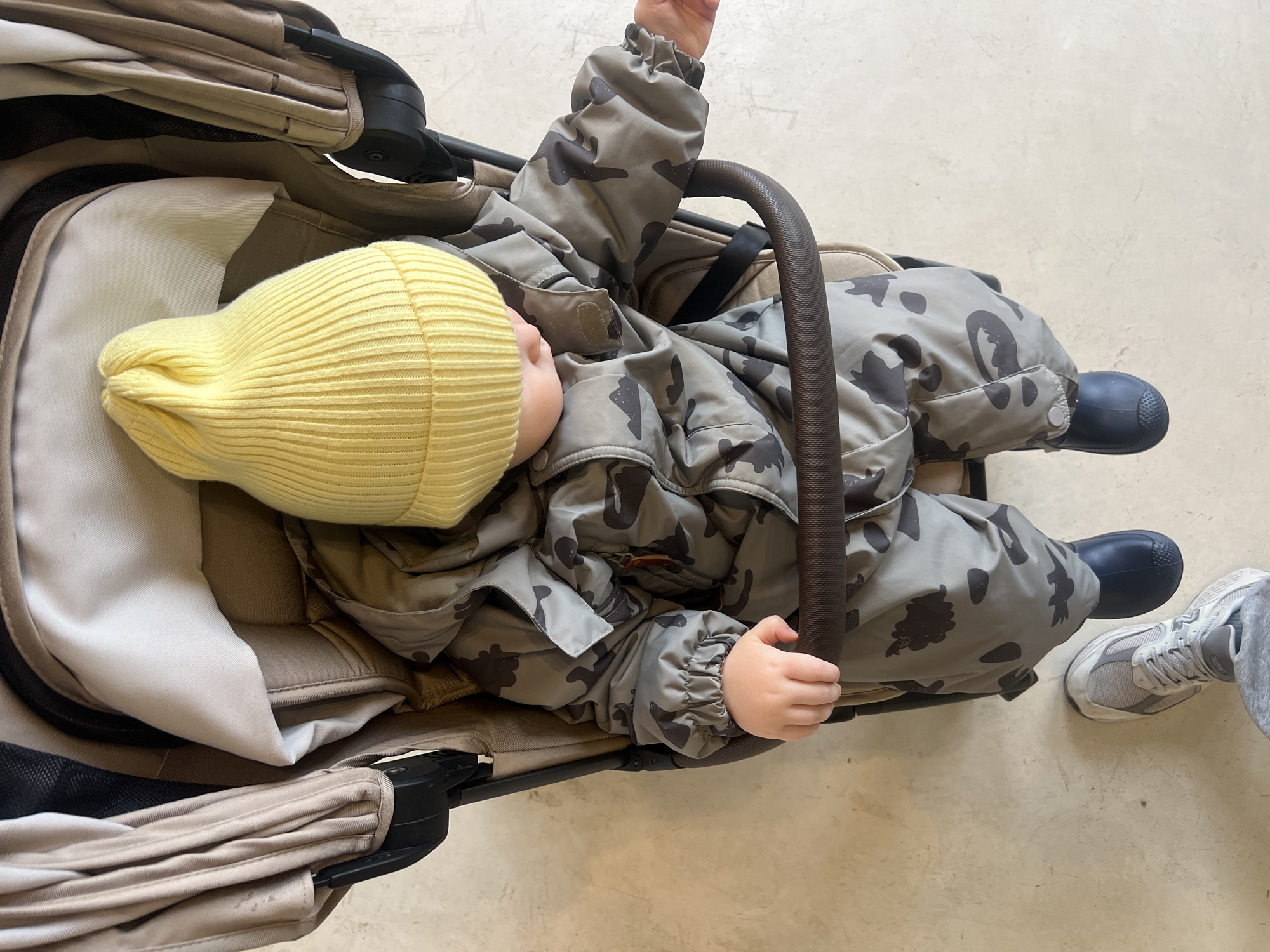 A top-down, overhead view captures a baby or toddler dressed in warm attire, seated in a beige and black stroller. The child wears a bright yellow, ribbed knitted beanie that covers their head, and a grey-green camouflage-patterned snowsuit or padded jacket. Their small hands are visible, with the left hand resting on the stroller's dark brown, textured bumper bar. Dark blue or black boots are visible on the child's feet.
The stroller features beige fabric upholstery, a black frame, and a dark brown bumper bar. The setting appears to be an indoor or sheltered outdoor space, characterized by a light-colored, possibly concrete or tiled floor with subtle texture. In the bottom right corner, an adult's foot, clad in a grey athletic shoe with white soles and grey sweatpants, is visible, indicating an adult is standing near or pushing the stroller.
The warm clothing suggests cool or cold weather, aligning with the provided context of Khimki, Russia. The lighting is bright and even, indicating daytime. No specific activity beyond the child being seated in the stroller is clearly depicted. There is no visible text in the image.