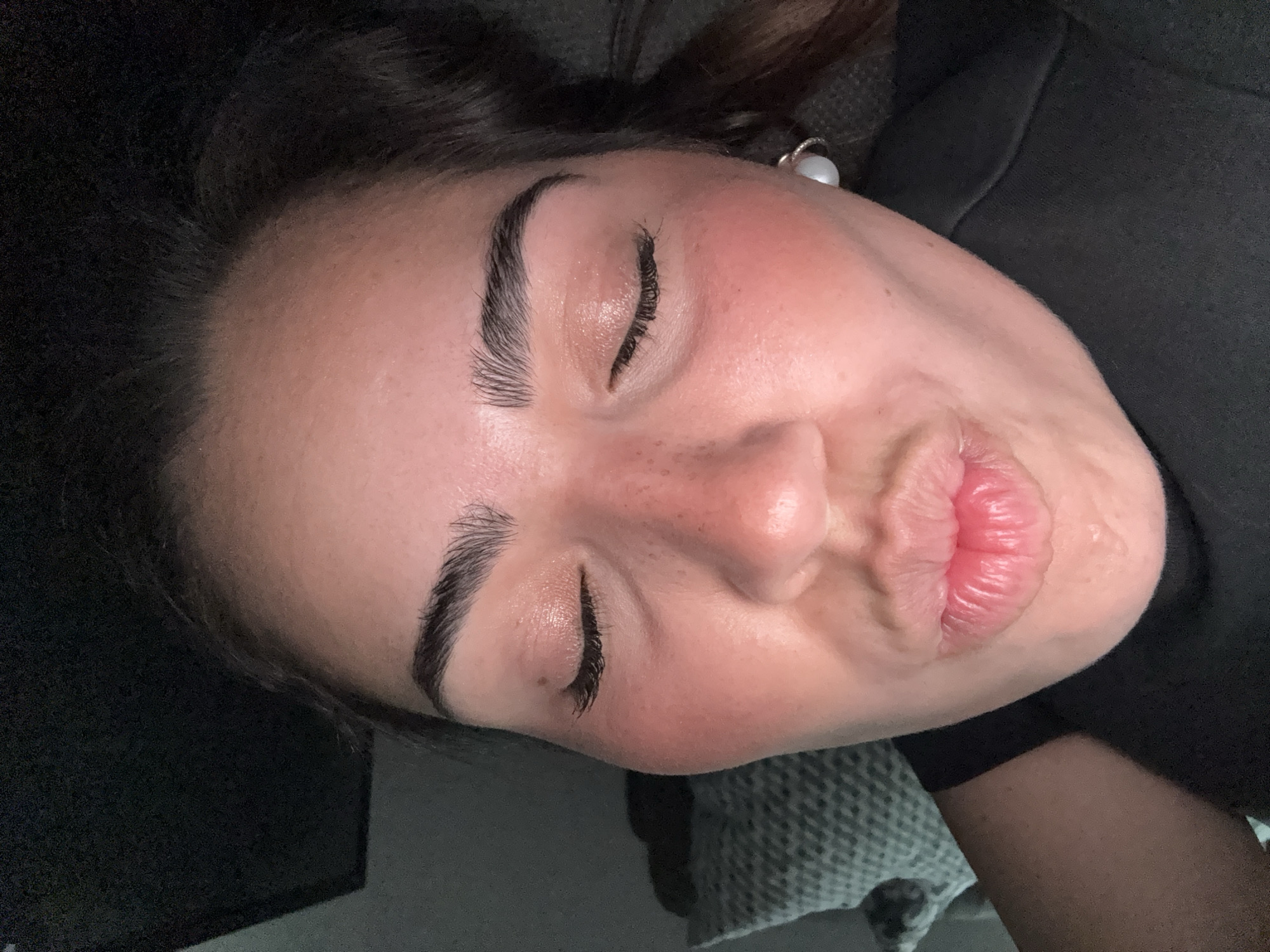 A person is lying down, facing the camera with their eyes closed and lips puckered. They have dark hair, defined eyebrows, and freckles. The skin on their face is reddish. Part of a dark-colored garment is visible. The location is unknown, it is NOT necessarily in the city of San Bernardino Tlaxcalancingo, Mexico.