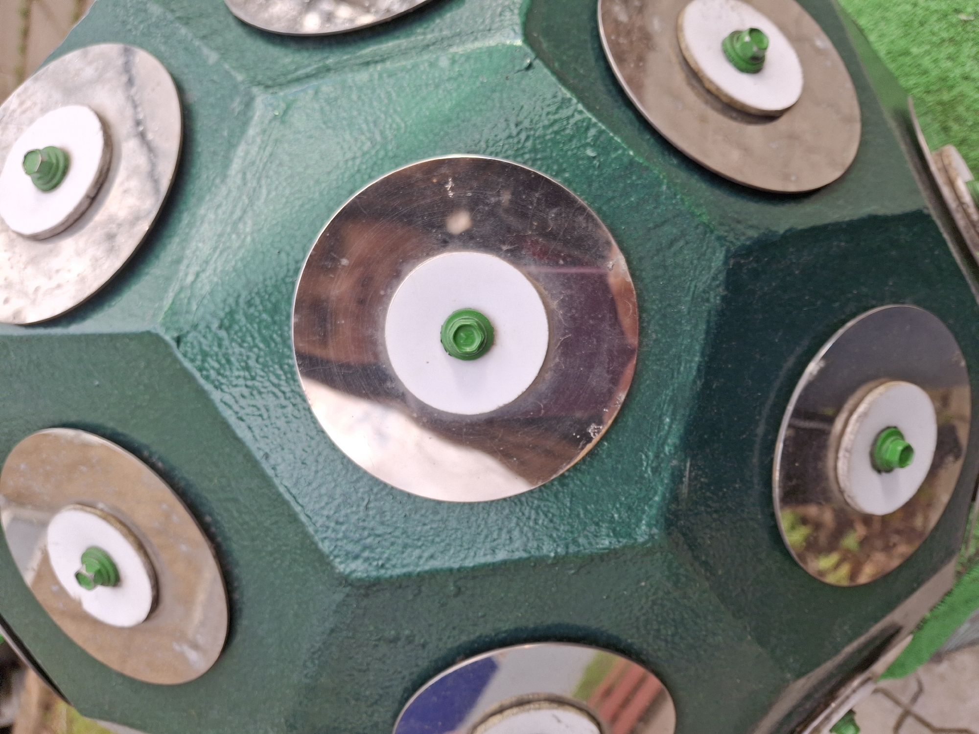 A close-up shot focuses on a textured, dark green polyhedral object, likely a sculpture or an interactive element, set outdoors in Sarapul, Russia. The object's surface is rough and uneven, consistent with a durable outdoor finish. Each visible face of the polyhedron features a layered circular adornment. These consist of a larger, reflective metallic disc as the base, topped by a smaller, matte white circular plastic disc. Centered on these layers is a small, bright green plastic fastener, possibly star-shaped or multi-pronged, securing the components to the green body.
Natural daylight illuminates the scene, casting reflections onto the metallic discs. These reflections include blurred green foliage, hinting at nearby trees or bushes, and in several instances, the clear reflection of a person holding a camera, suggesting the object is at an accessible height. Some metallic discs show minor scratches, smudges, and variations in reflection, including hints of other surrounding elements like muted reddish-brown or bluish structures. The overall impression is one of a robust, possibly artistic or recreational installation designed for a public space. No specific activity is depicted, and no text is visible.