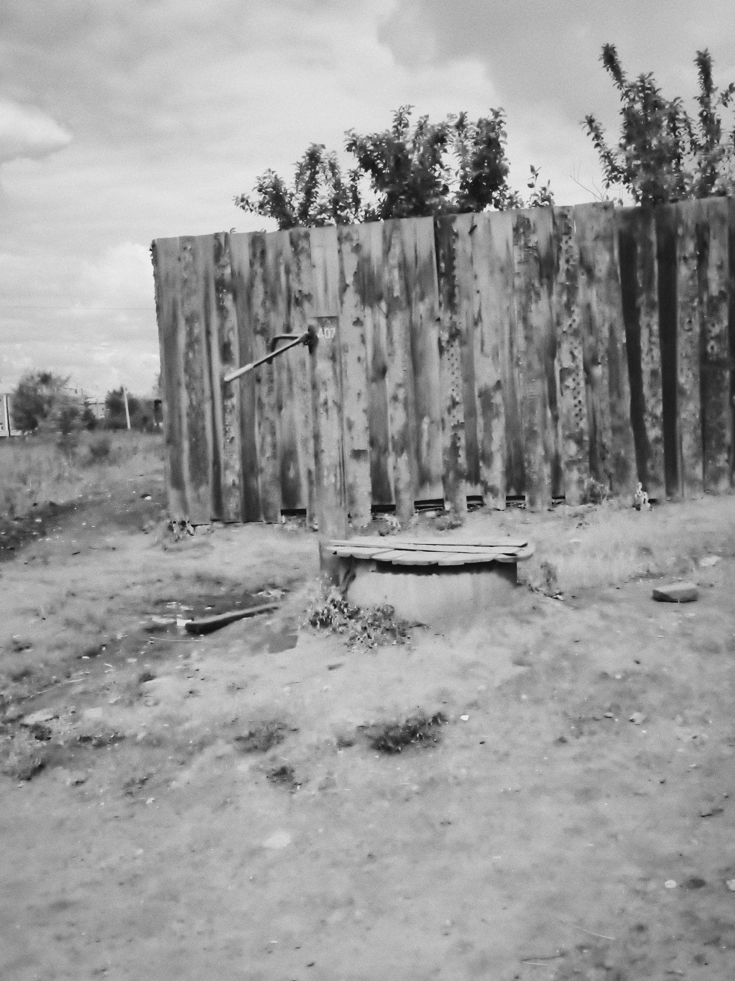 The black and white image depicts an outdoor scene in Cheremkhovo, Russia, featuring a corrugated metal fence and a well. The fence is weathered and appears to be a significant structure in the area, with some trees and vegetation growing behind it. In front of the fence, a circular well with a wooden cover is visible. The ground is uneven and covered with grass and dirt, suggesting an undeveloped or rural area within the city. The sky is cloudy, indicating a possibly overcast day. There are no people or animals clearly visible in the image. The overall impression is one of a simple, utilitarian environment, possibly a backyard or a less-maintained part of the city.