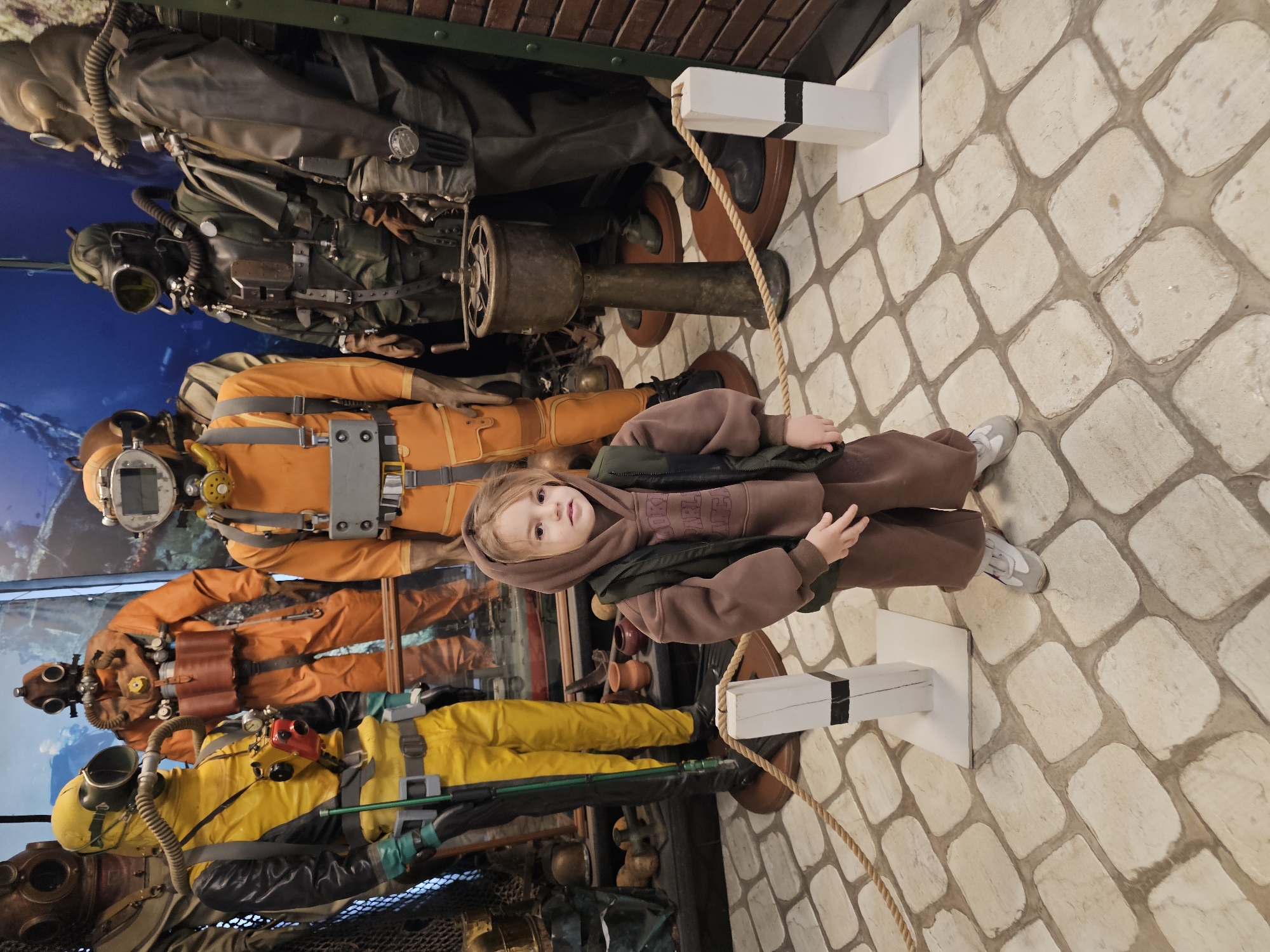 A child stands in front of a display of diving suits at what appears to be the museum or exhibition in Архангельское, Russia. The child wears a brown hoodie and matching pants with a dark vest and light gray shoes. They are standing on a light-colored, cobblestone-patterned floor. Behind the child is a multi-level display of vintage diving suits in various colors, including yellow, orange, and black. The diving suits are complete with helmets and equipment. The backdrop behind the suits appears to be a mural or a photograph depicting an underwater scene. A rope barrier is present in front of the diving suits, and the suits are mounted on wooden bases. A brick-patterned wall is visible at the top of the frame.