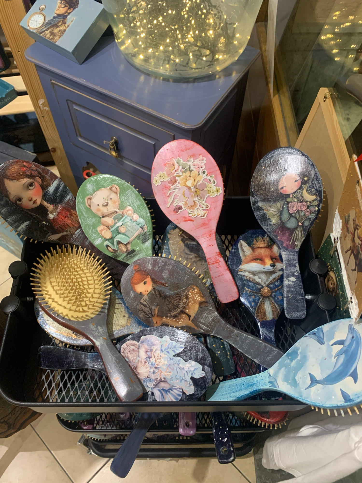The image displays a collection of vintage-style hairbrushes, each adorned with a unique illustration. The brushes are arranged in a black wire basket, which appears to be on display in a shop. Some of the illustrations depict whimsical characters such as a girl with large eyes, a teddy bear in a car, a fox wearing a crown, and a girl holding flowers under a starry sky. Other brushes feature floral designs and a pair of dolphins swimming in the sky. In the background, a blue drawer cabinet is visible, topped with a glass vase filled with fairy lights. A wooden ladder-like structure is also present, adding to the rustic aesthetic of the display. The overall impression is that of a curated collection of decorative items, likely found in a boutique or antique shop. The scene is set in Skopelos, Greece.