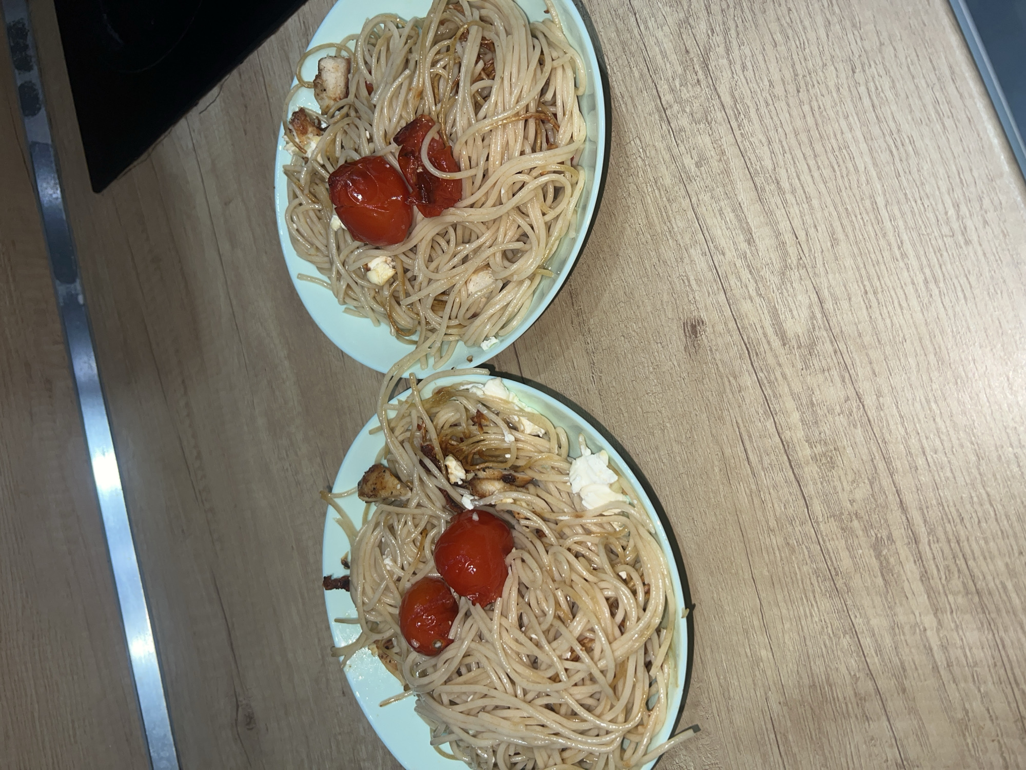 The image shows two plates of spaghetti with tomatoes, cheese, and what appears to be chicken or tofu on a light brown wooden surface. The plates are light blue. There are metallic elements partially visible on the left edge. There is no information to indicate the location is Plovdiv, Bulgaria.