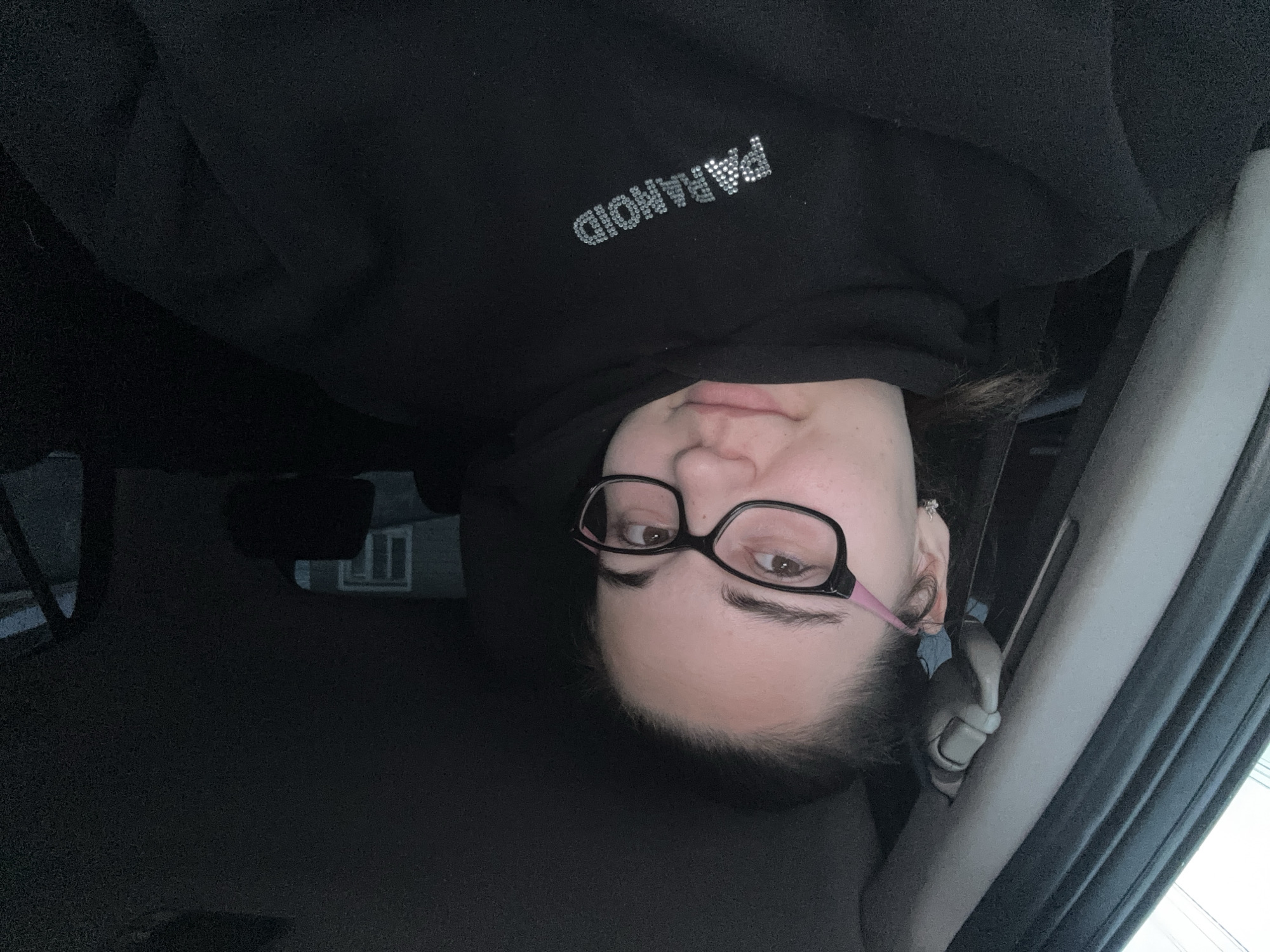 The image shows a woman wearing glasses and a black hoodie with the word "PARANOID" spelled out in rhinestones. She appears to be inside a car, possibly in the driver's seat or passenger seat. The interior of the car is dark. There is a window on the right, and a glimpse of a building can be seen through the windshield in the background. The location is not identifiable as Zlatitsa, Bulgaria from the given details.