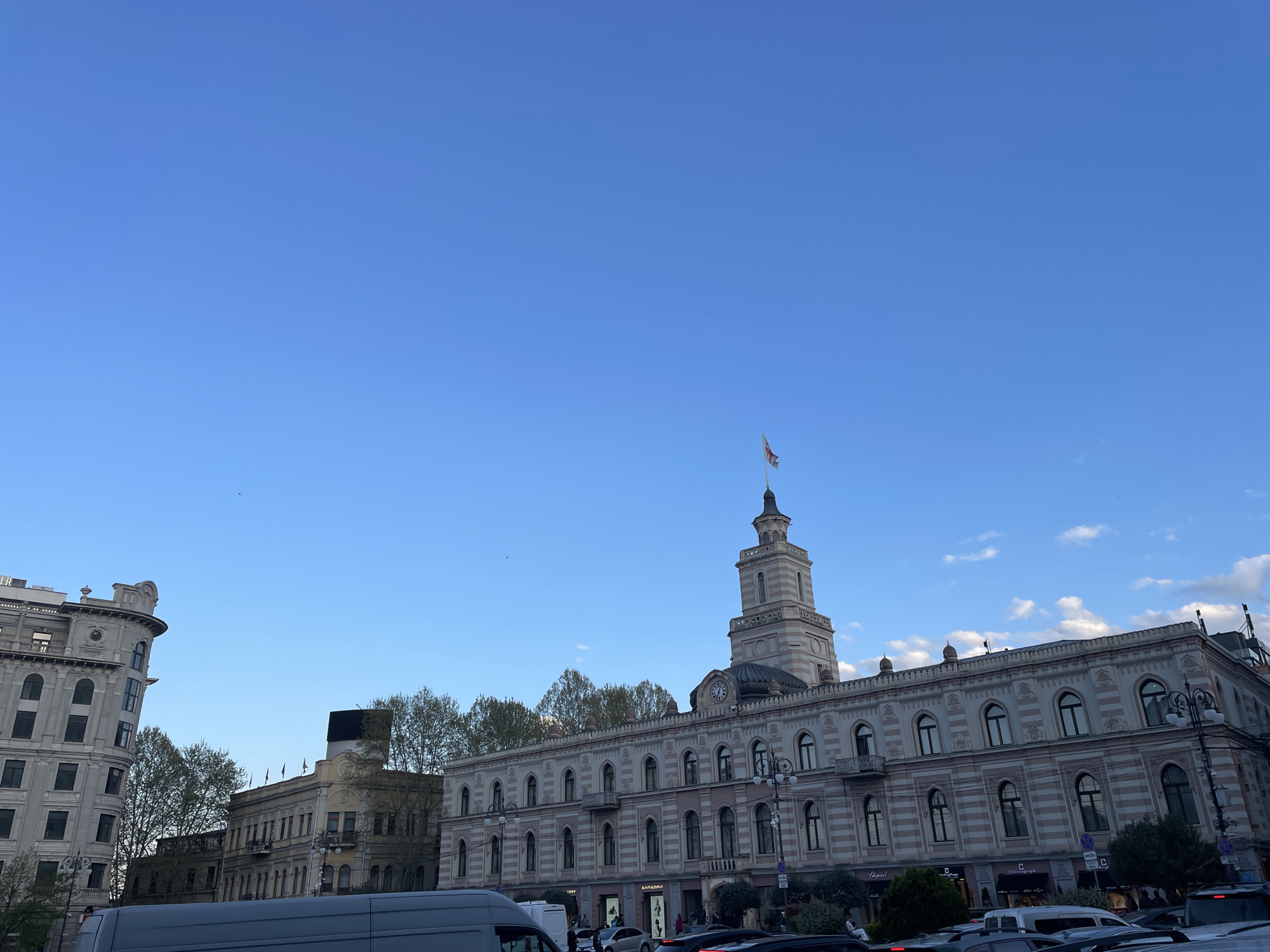 This image captures a daytime urban street scene in Tbilisi, Georgia, under a clear, bright blue sky with a few scattered white clouds. The central feature is a majestic, multi-story building characterized by an ornate facade with alternating light and darker striped stone patterns, numerous arched windows, and decorative balconies. A prominent clock tower rises from the building's roof, topped with a spire from which the Georgian national flag is flying.
To the left, another large, architecturally distinct building with rounded sections and many windows is partially visible, contributing to the historic city aesthetic. Further back, several other buildings and green-leafed trees can be seen, hinting at a tree-lined street or square.
The foreground reveals a busy street with various vehicles, including a white van in the immediate foreground and multiple cars forming part of the traffic flow. Pedestrians are visible on the sidewalks, particularly in front of the ground-level shops of the main building.
Visible text on the shopfronts includes "CHOPARD" and "Chronograph," indicating luxury retailers. Some Georgian script, possibly "მამაჩემი" (Mamachemi - My Father), can also be discerned on a sign. The bright, even lighting suggests it is daytime, possibly mid-morning or early afternoon, with pleasant, clear weather conditions. The scene portrays a bustling city center with significant architectural heritage.
